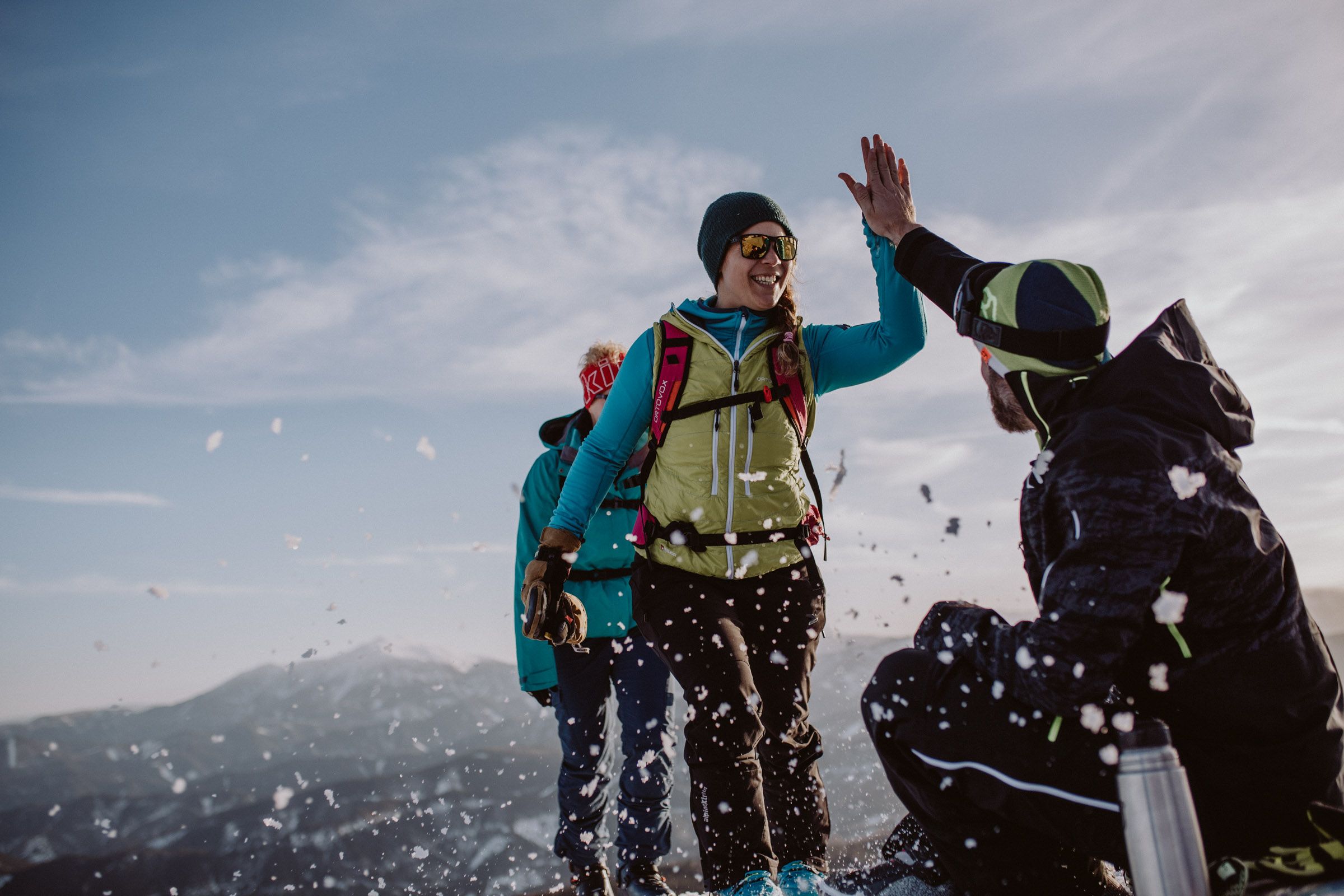 Three people in the snow, one high-fiving another, with mountains in the background.