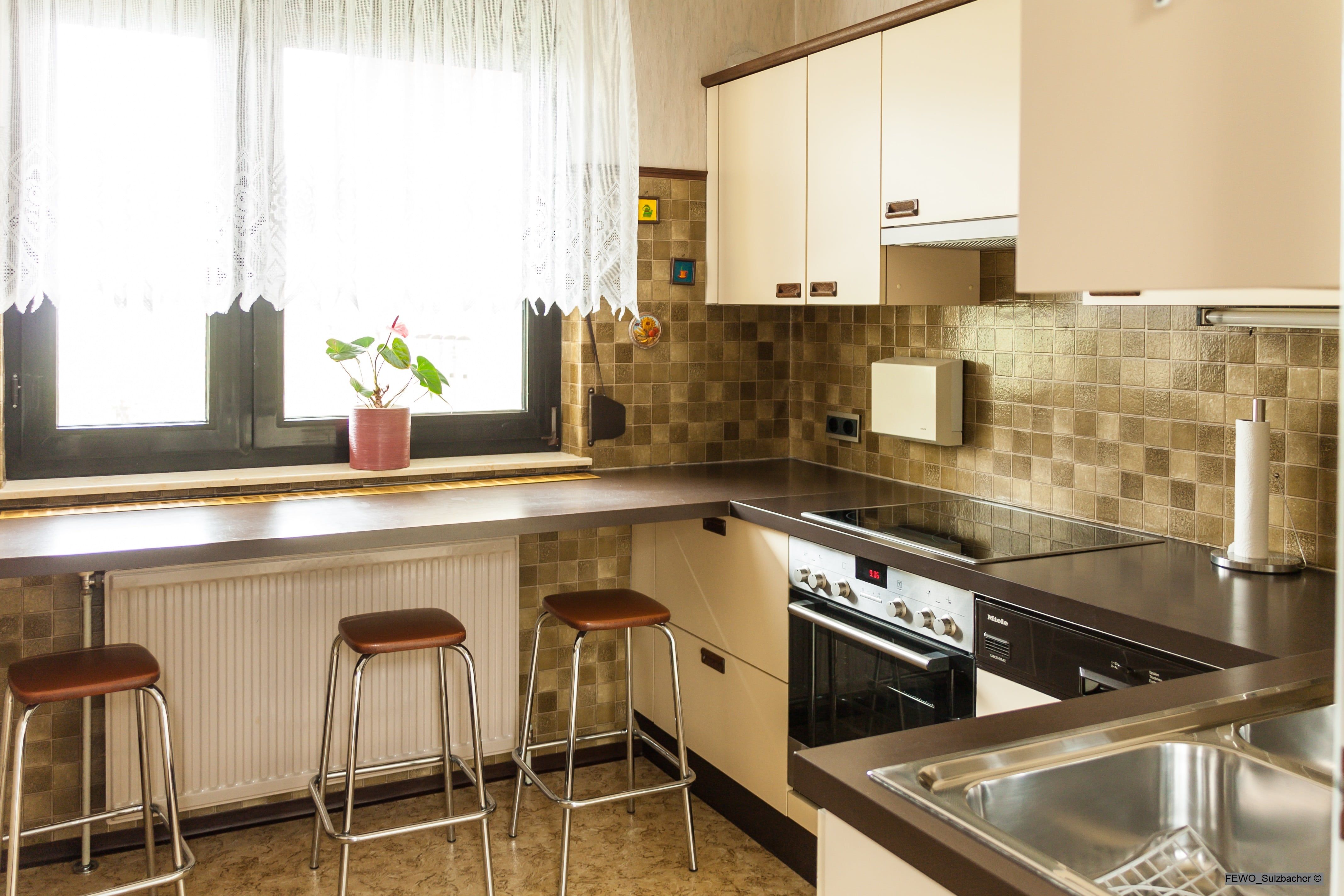 Modern kitchen with brown tiles, stove, sink and bar stools in front of a window with curtains.