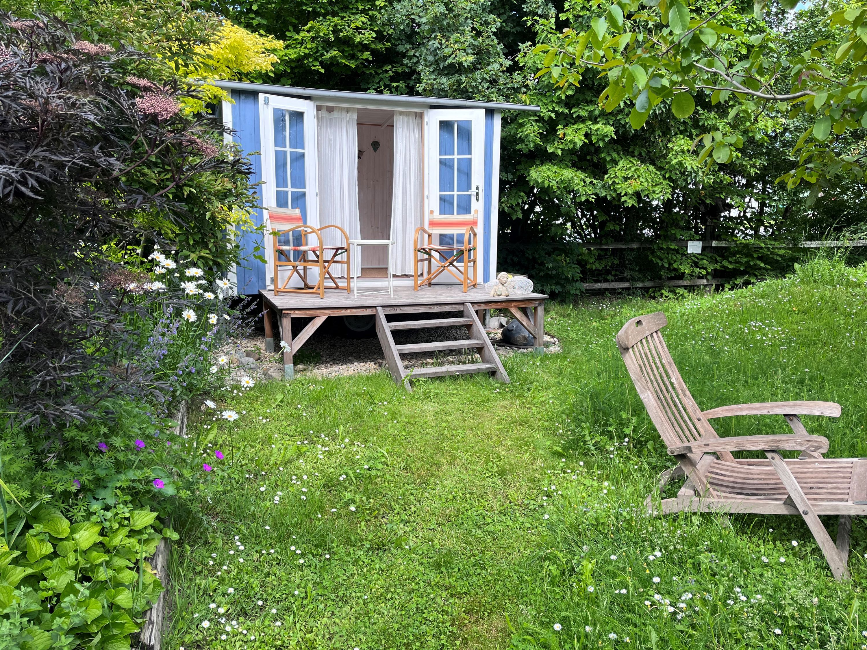 A blue garden pavilion with two chairs on a wooden veranda, surrounded by a green meadow and colorful flowers.