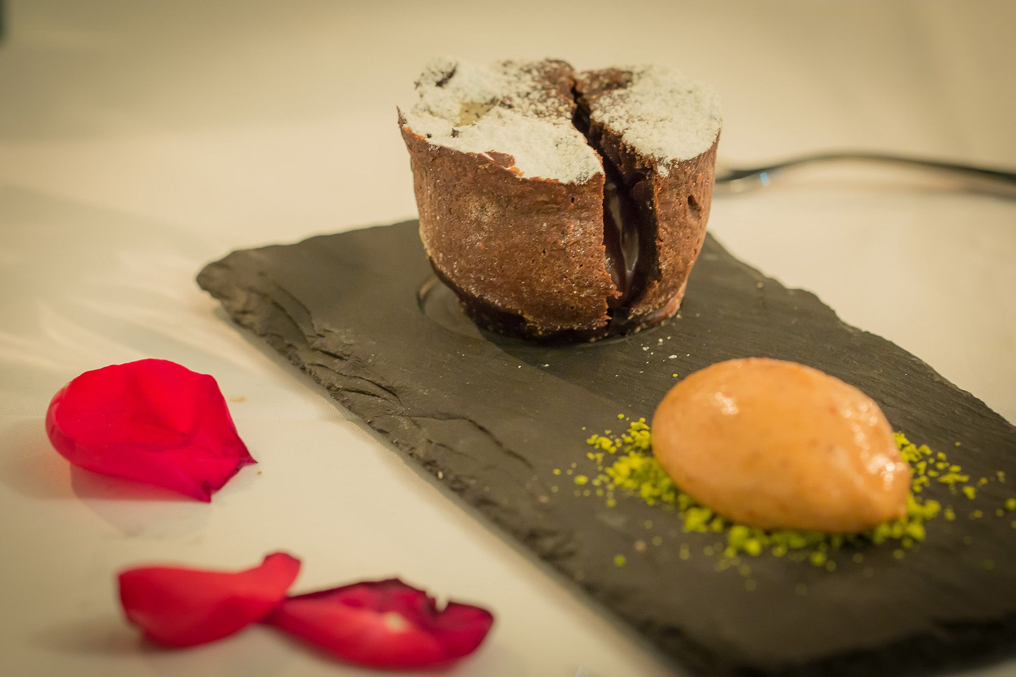A chocolate volcano dessert on a slate plate with a scoop of ice cream and red rose petals.