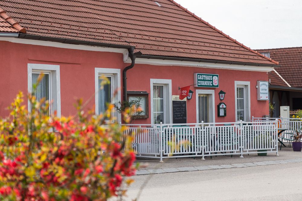 A pink building with the inscription 'Gasthaus Zivanovic', surrounded by a white fence and flowers in the foreground.