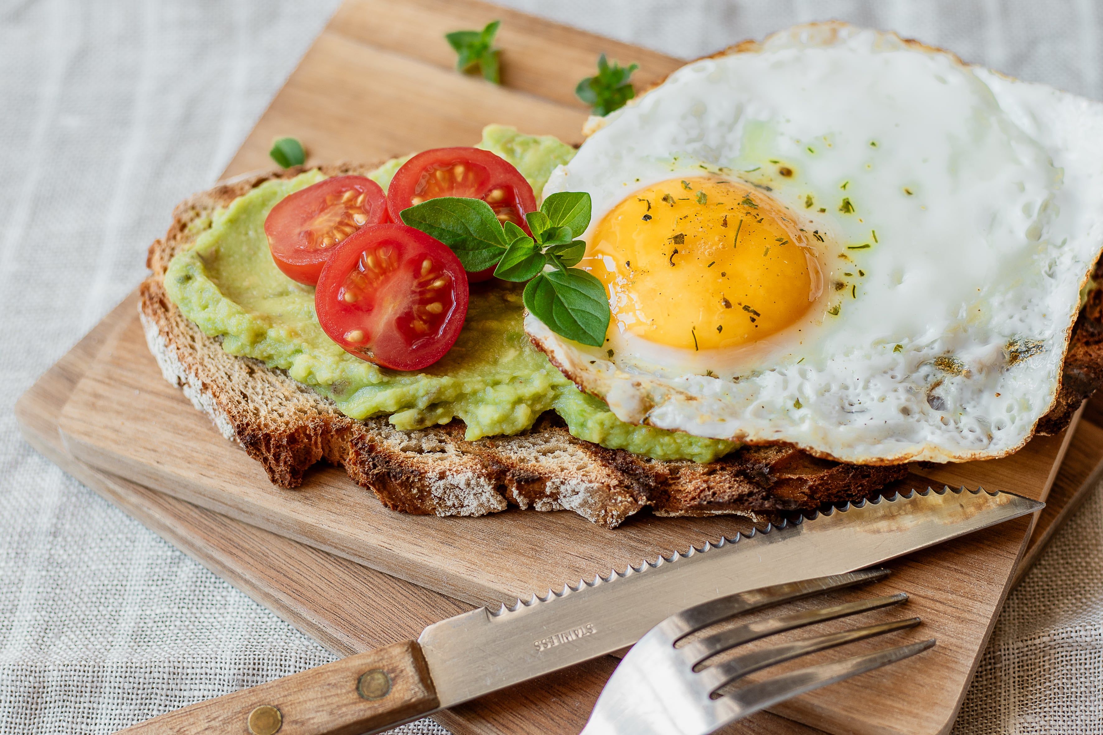 A wooden board with a slice of bread, topped with avocado cream, tomato slices and a fried egg.