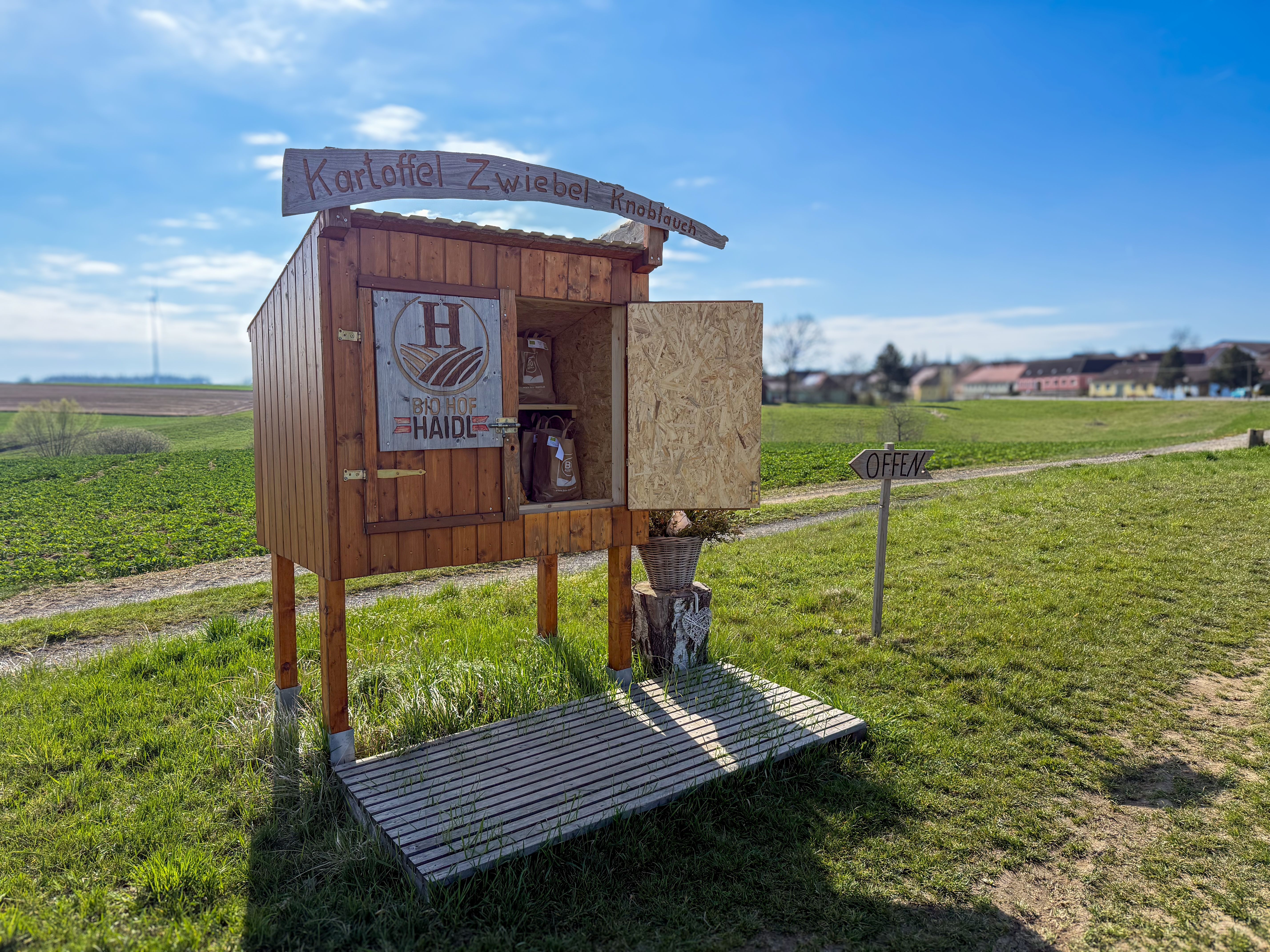 A small stall on a country lane with the inscription 'Kartoffel Zwiebel Knoblauch' and 'Bio Hof Haide'. A sign says 'Open'.