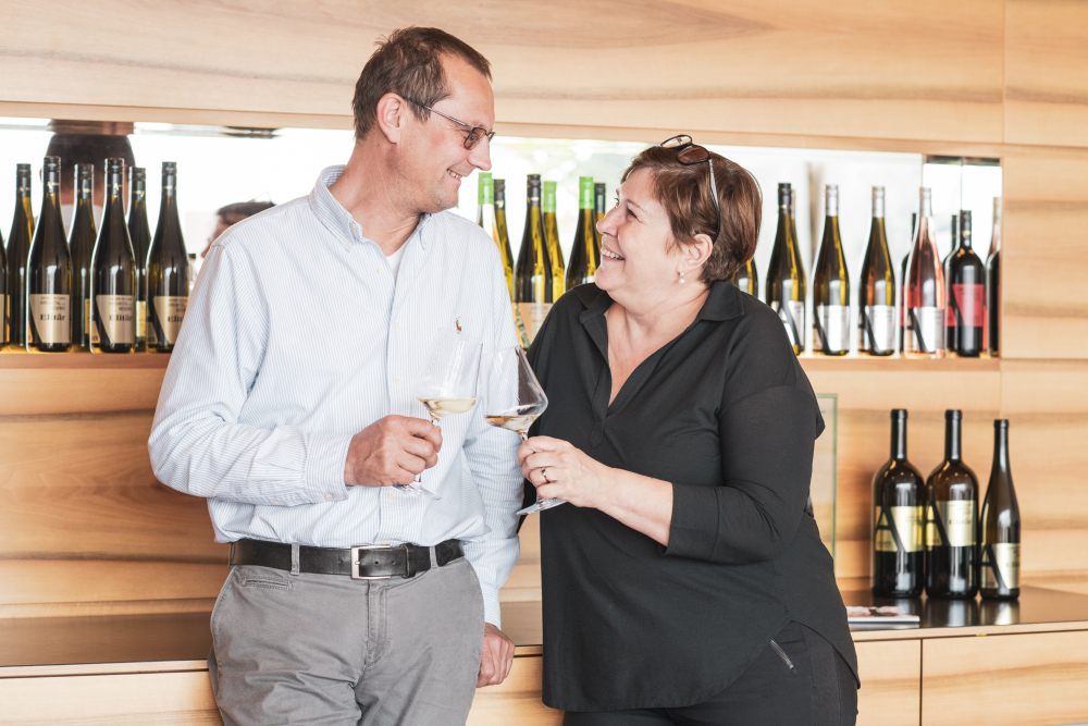 A man and a woman toast with wine glasses, wine bottles in the background.