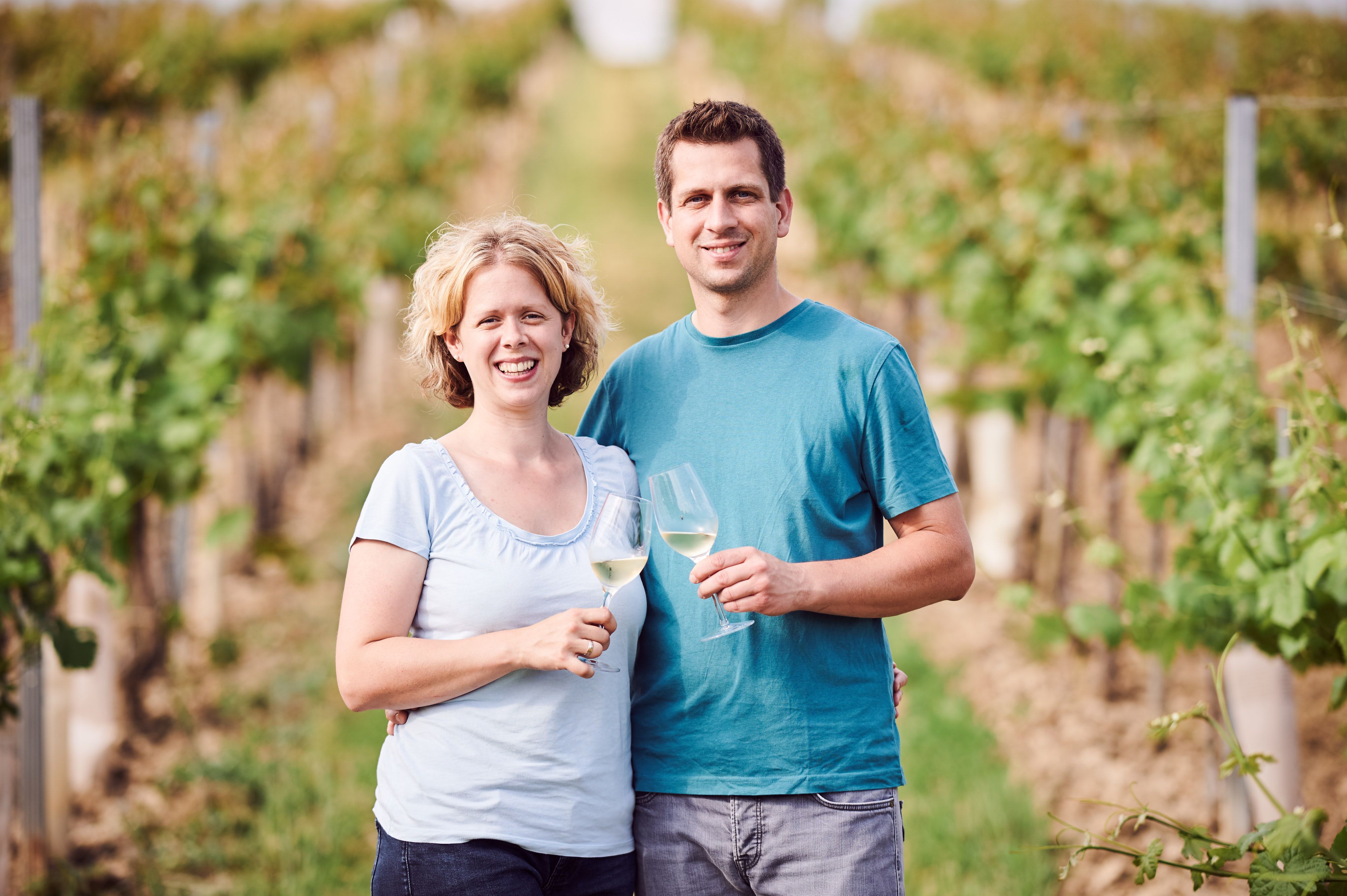 A smiling couple stands in a vineyard holding glasses of wine.
