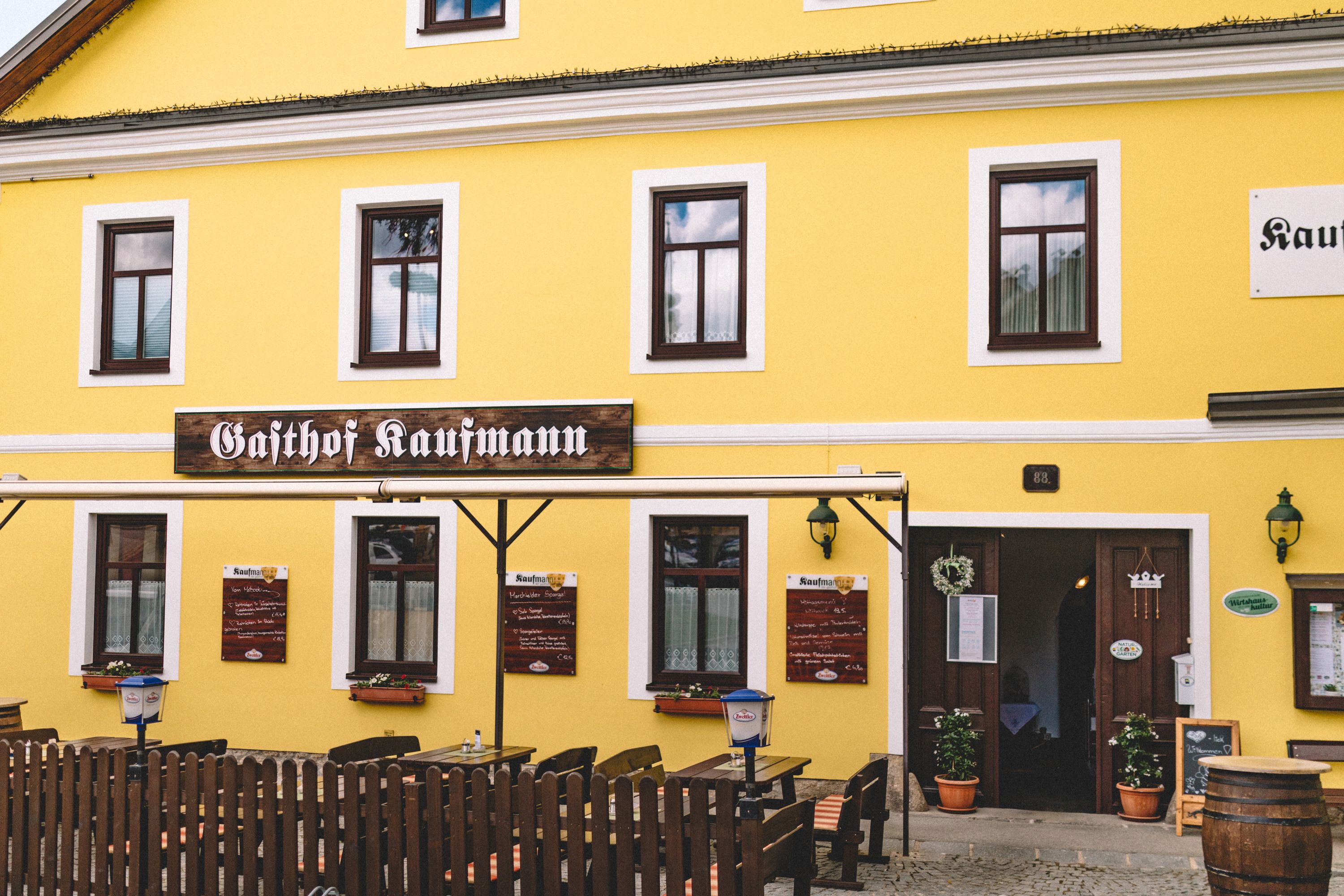 Yellow building with the inscription 'Gasthof Kaufmann', wooden tables and chairs outside.