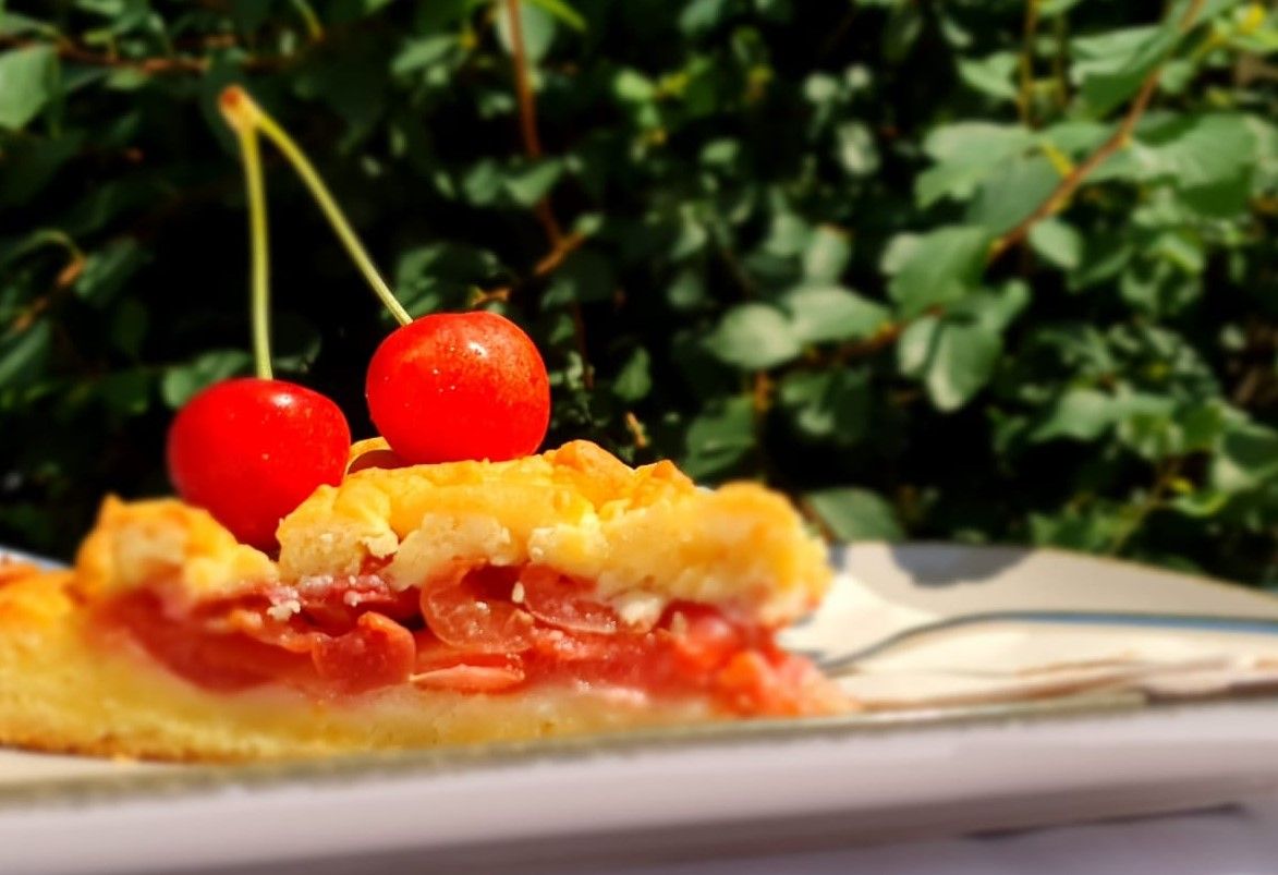 A slice of cherry cake with two cherries as decoration, green leaves in the background.