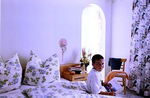 A boy sits on a bed in a bright room with floral curtains and bed linen.