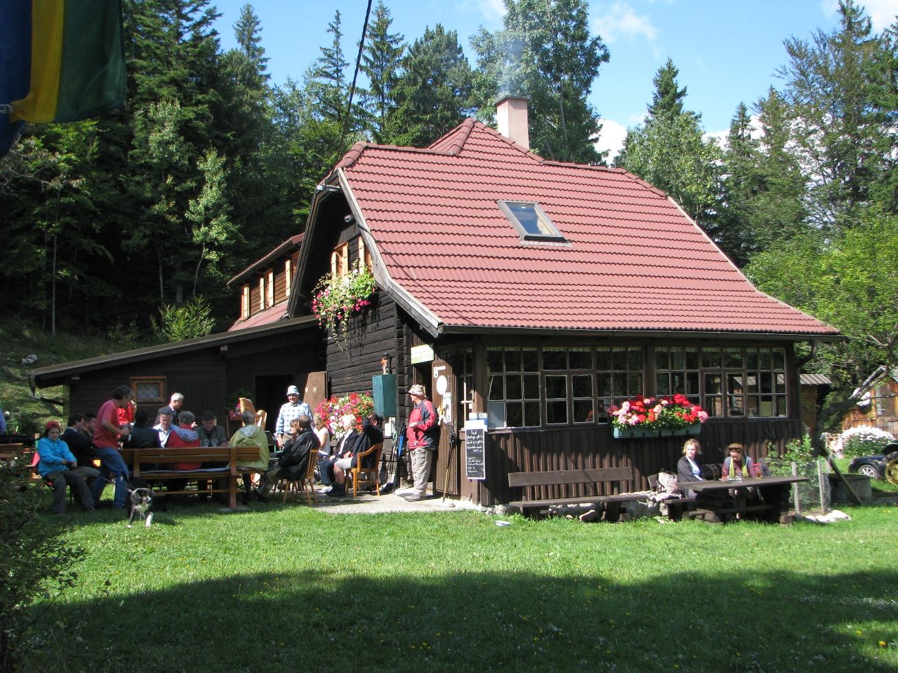 A mountain hut with a red roof, surrounded by forest, with people sitting outside.