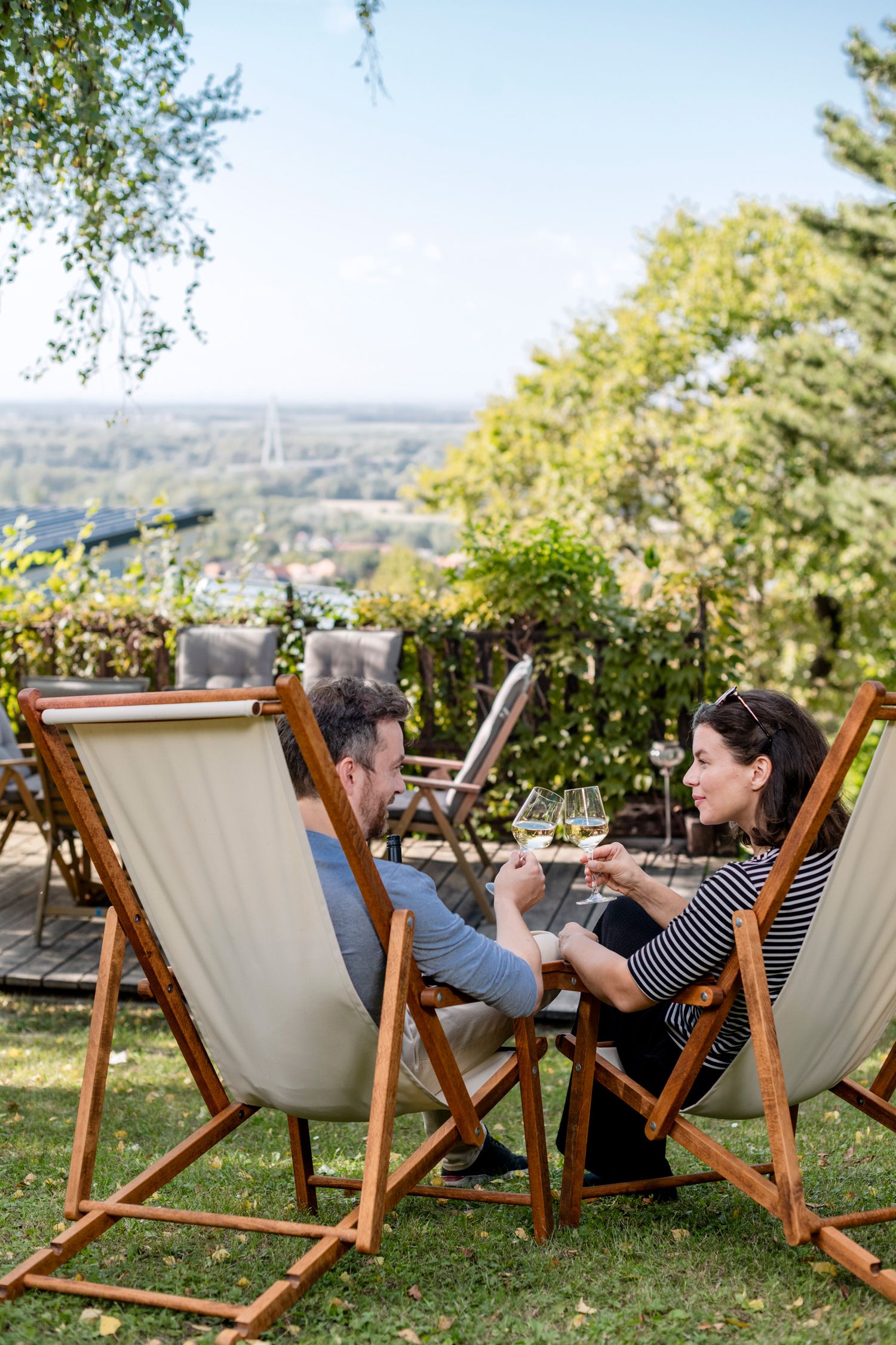 A couple sits in deckchairs in the garden and toasts with glasses of wine, overlooking a green landscape.