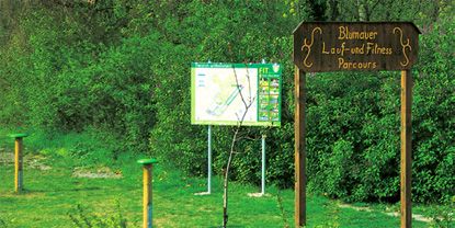 Wooden sign with the inscription 'Blumauer Lauf- und Fitness Parcours' in front of an information board in the countryside.