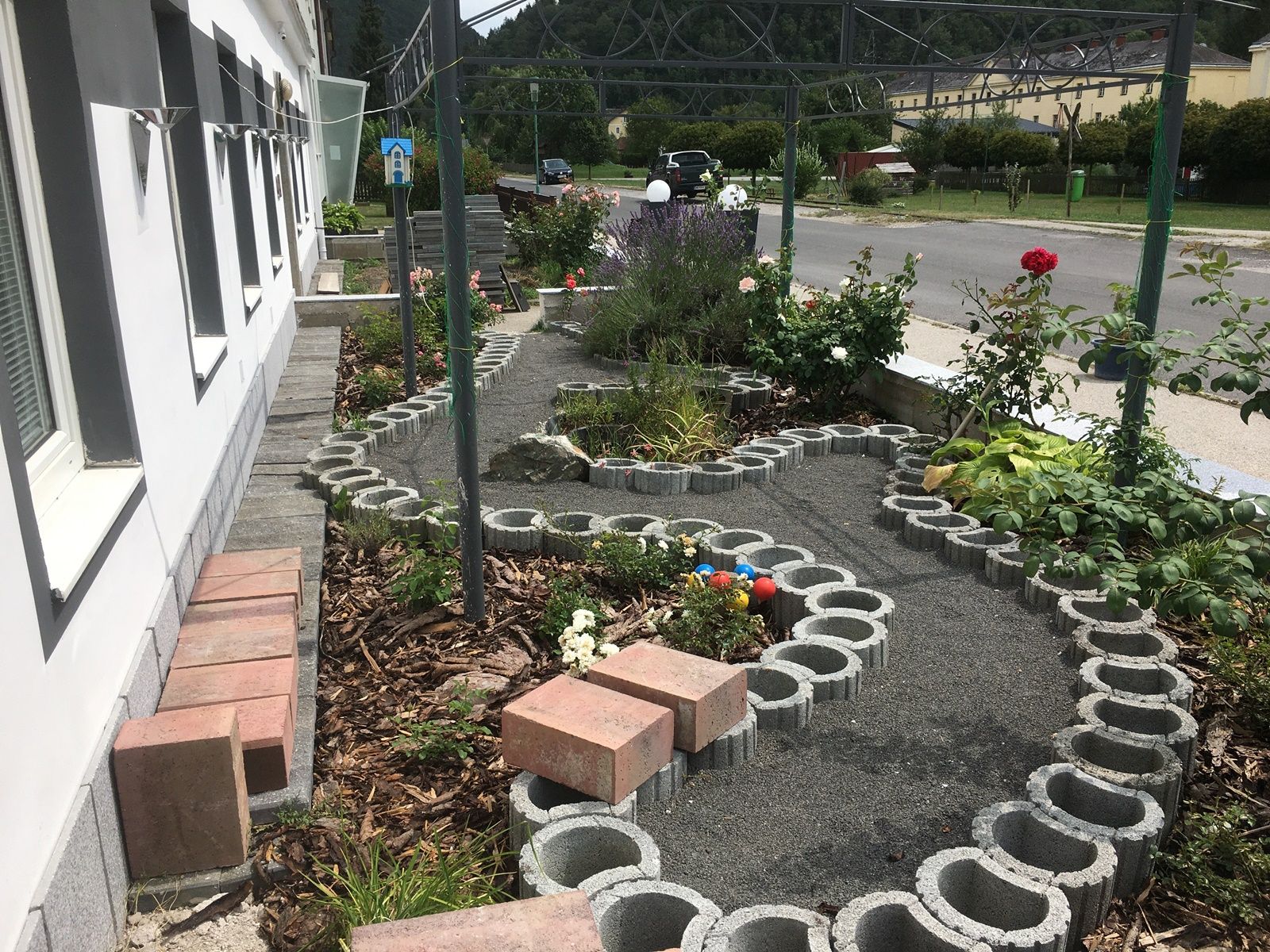 A small garden with roses and a curved path made of concrete blocks next to a building.