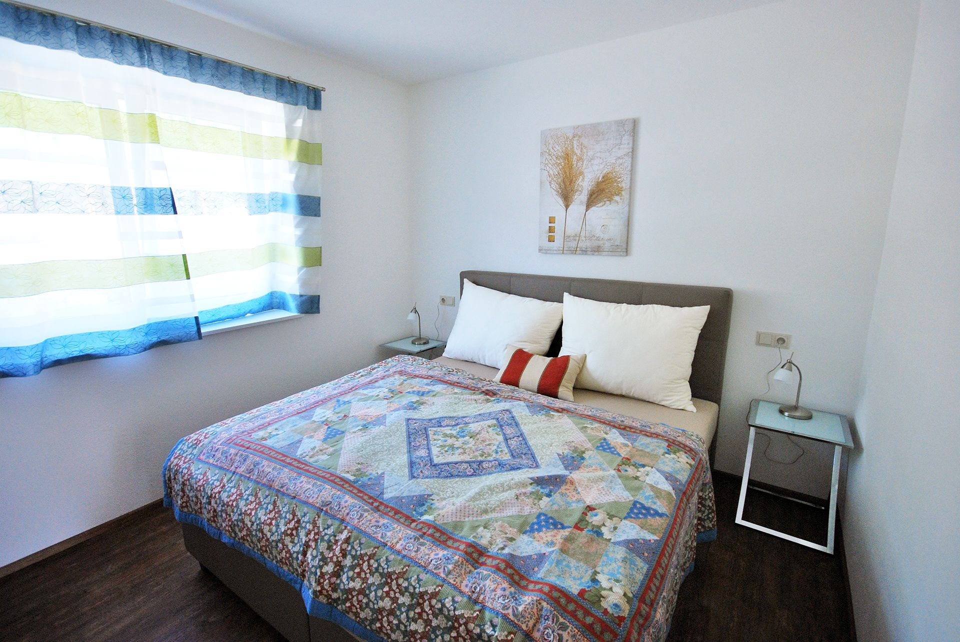 A bedroom with a double bed, colorful bedspread, two bedside tables and a window with striped curtains.