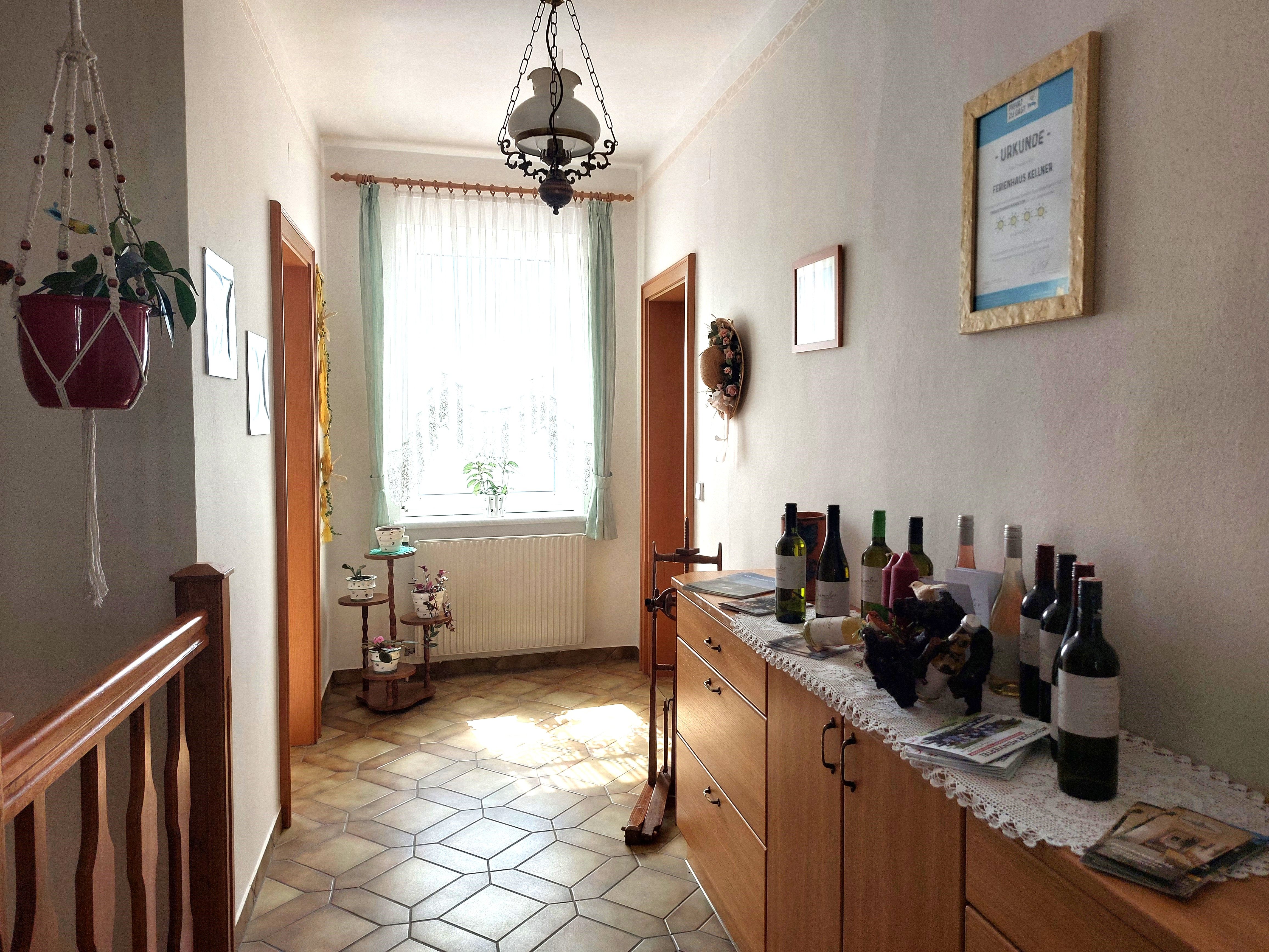 A bright anteroom with tiled floor, a chest of drawers with wine bottles and a window with curtains.