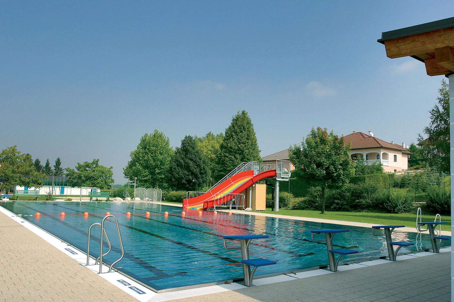 An outdoor pool with a swimming pool, a red slide and diving boards. Trees and a house can be seen in the background.