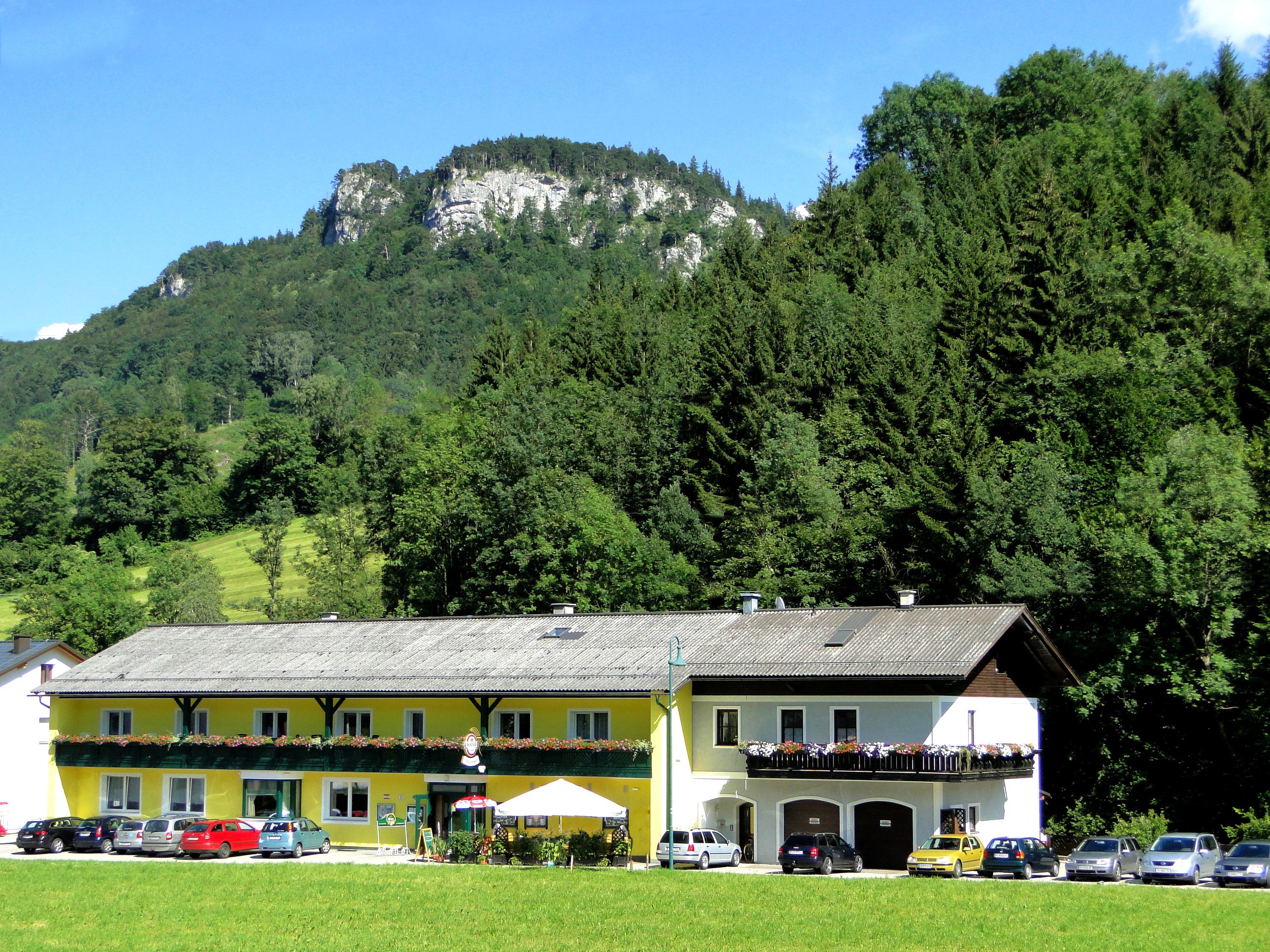 A yellow building with a balcony and flowers, surrounded by trees and a mountain in the background.