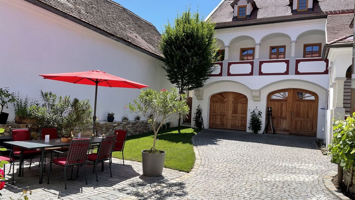 An inner courtyard with a red parasol, table and chairs, surrounded by plants and a building with wooden doors and windows.
