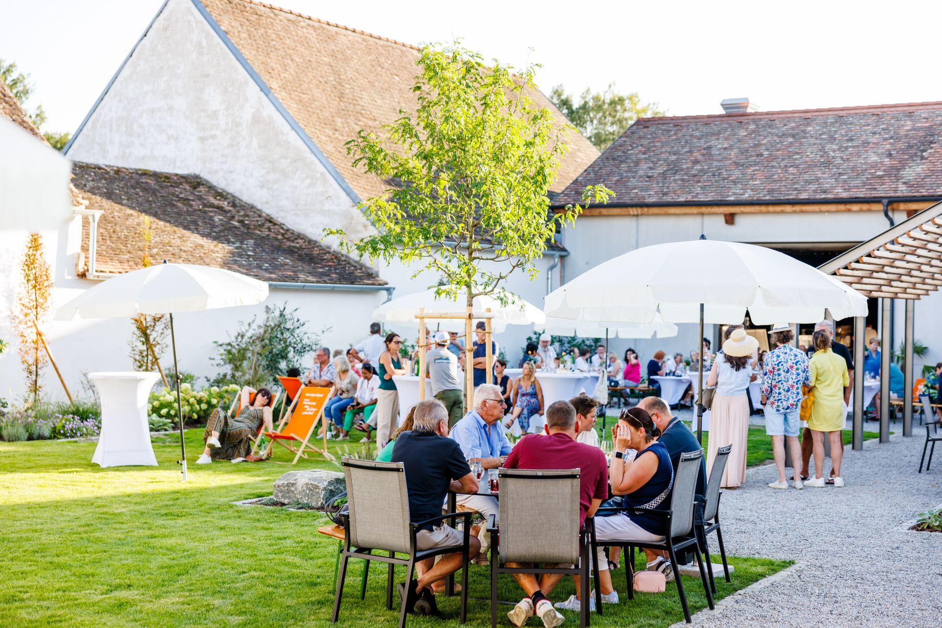 People enjoying an outdoor event with tables, chairs and parasols on a lawn in front of a building.