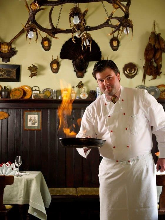 A chef in a white uniform flambées a dish in a rustic kitchen with hunting trophies on the wall.