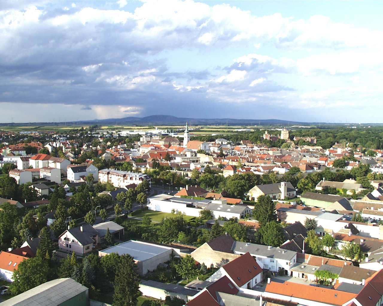 Aerial view of Bruck an der Leitha with houses and church, surrounded by green landscape and cloudy sky.