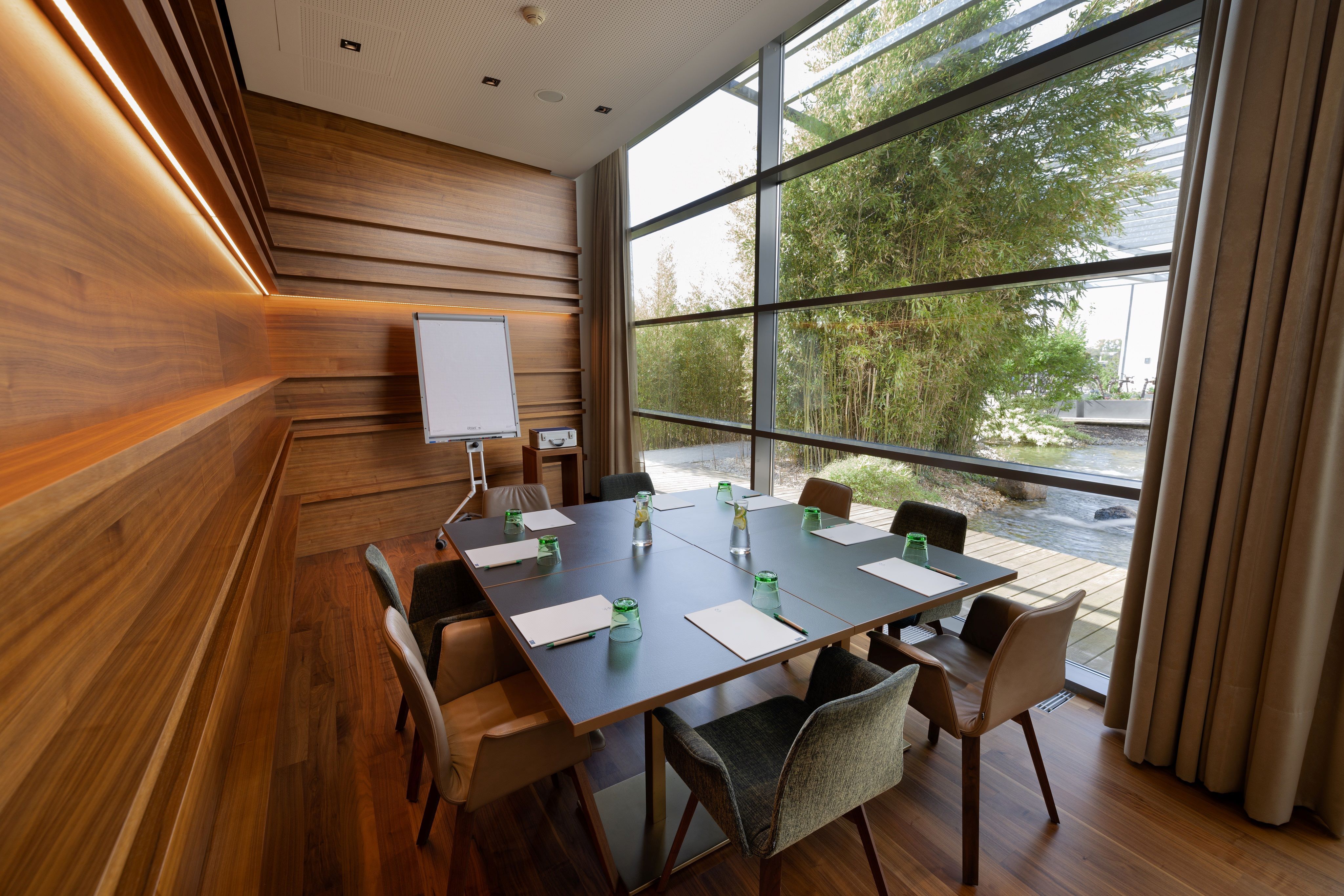 Modern seminar room with wooden walls, large table, chairs and flipchart. Large windows with a view of the garden.