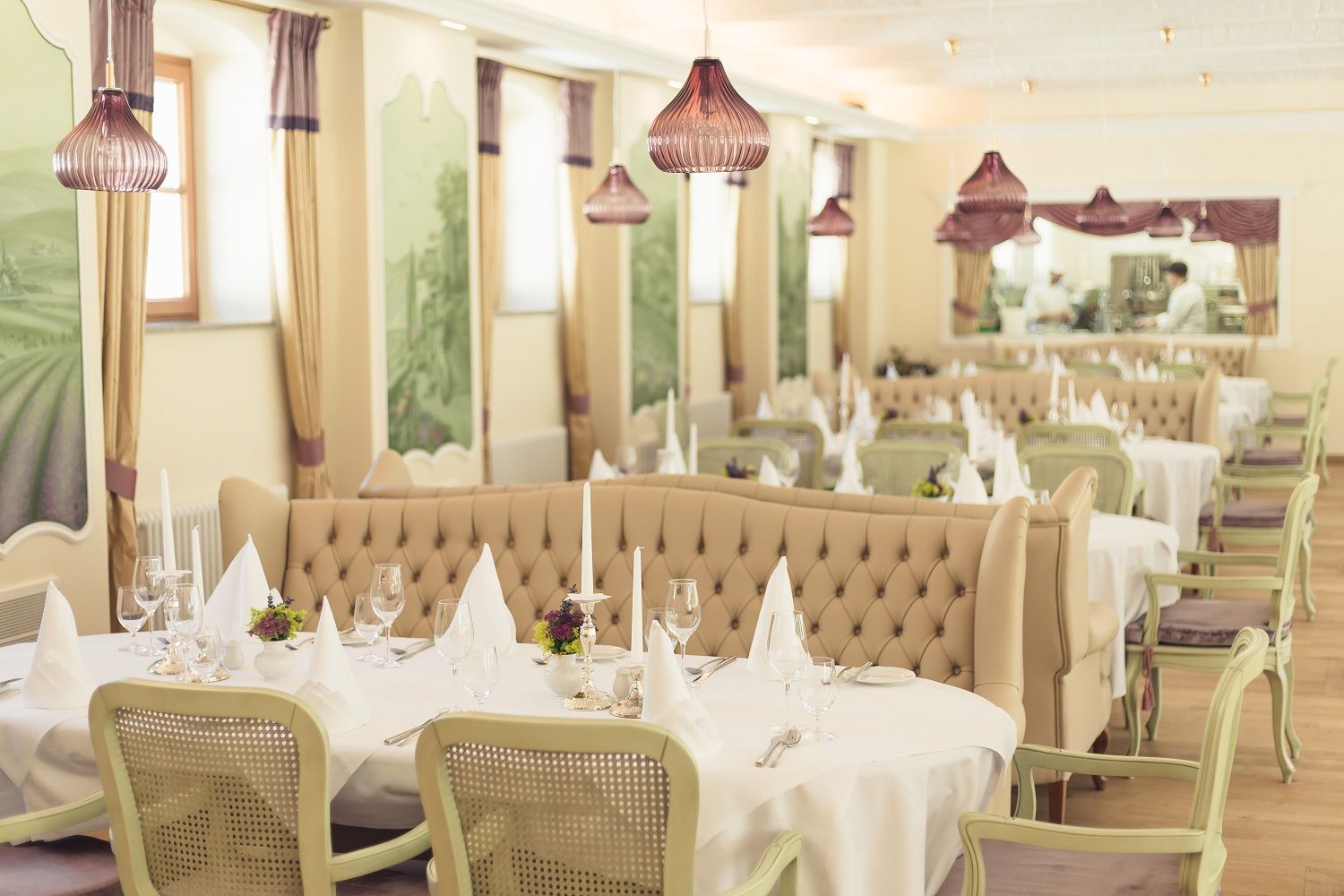 Elegant restaurant with laid tables, white tablecloths and candlesticks, beige upholstered benches and green chairs, decorative murals.