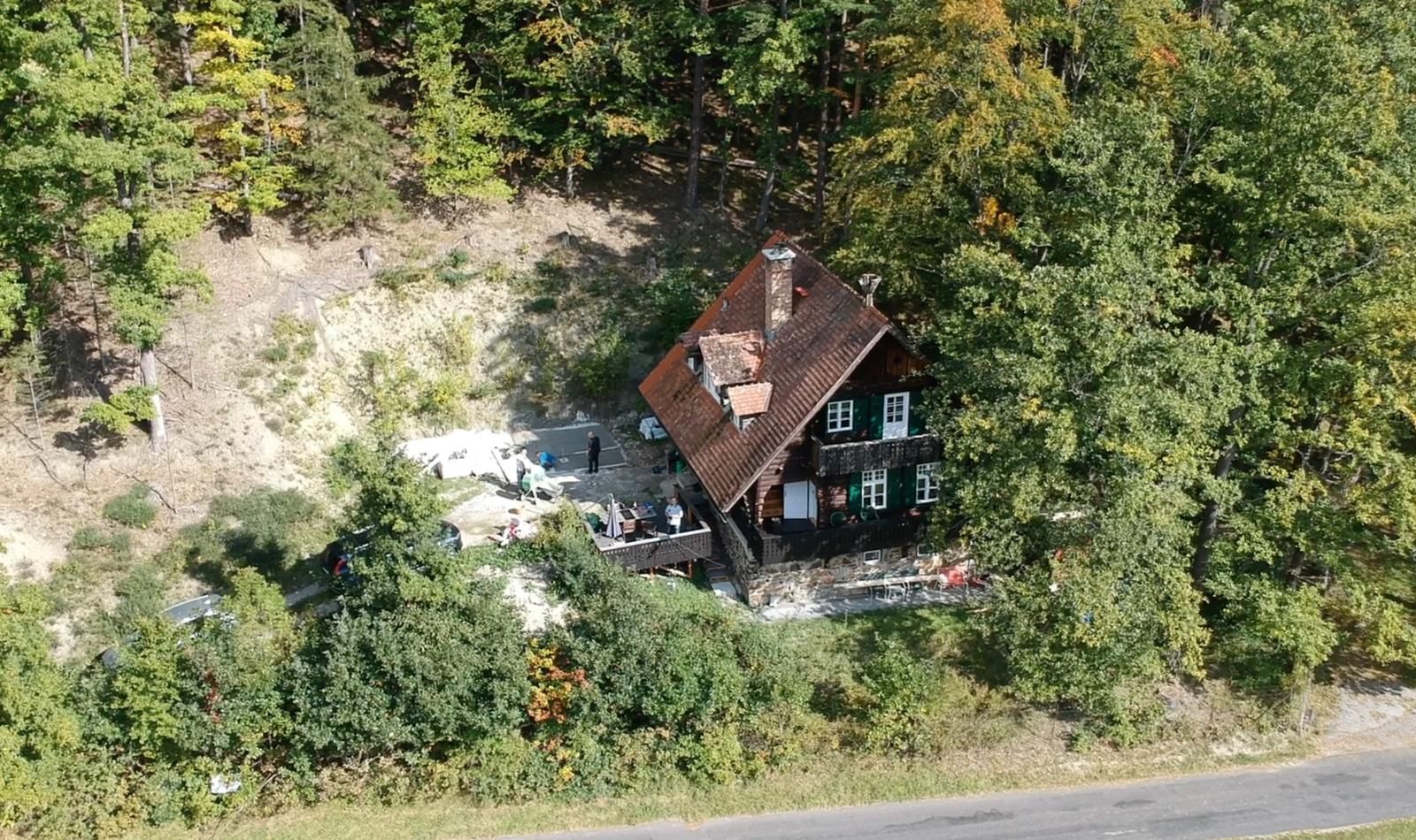 Aerial view of a house in the forest with a red roof and green shutters, surrounded by trees.