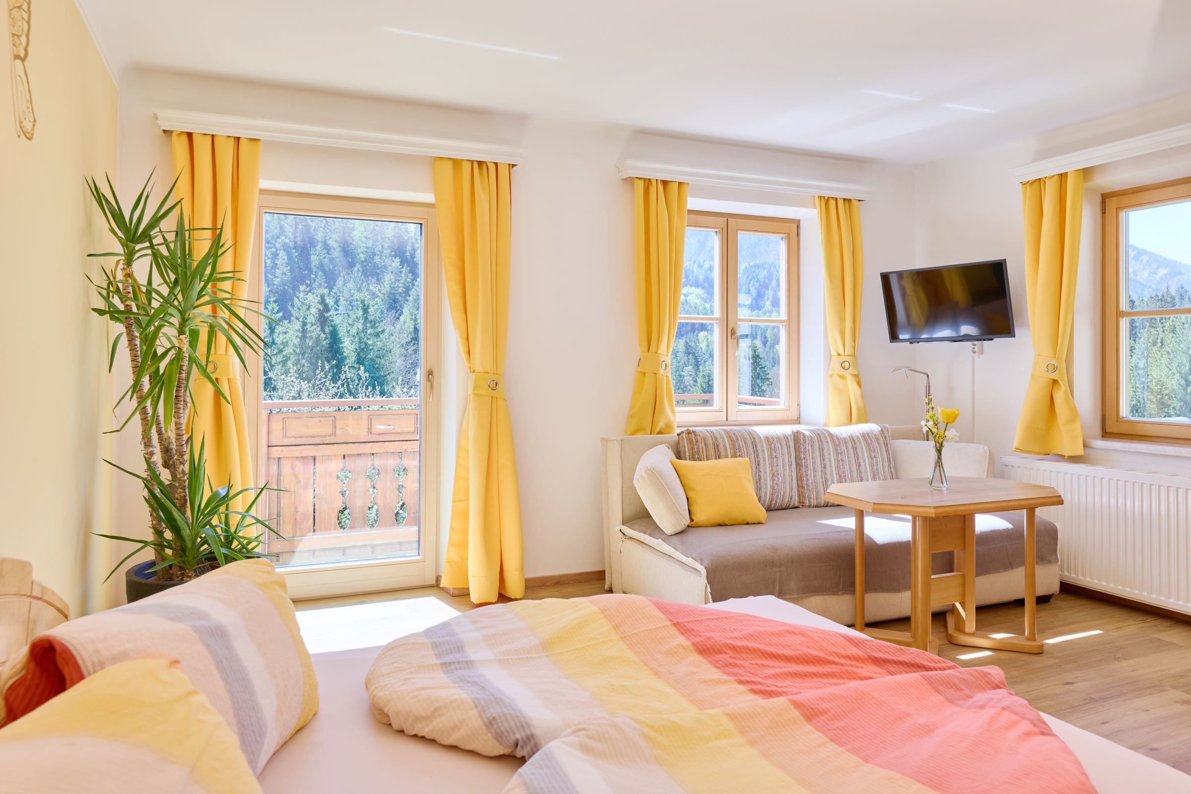 Bright room with bed, sofa, table and yellow curtains, view of the forest and mountains.