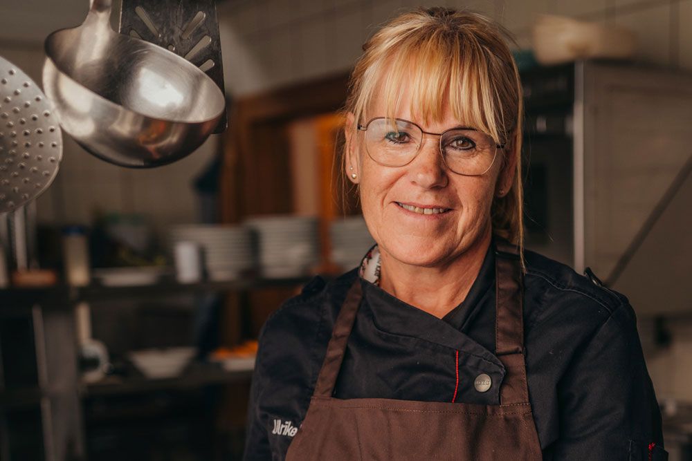 A woman in chef's clothes stands in a kitchen and smiles at the camera.
