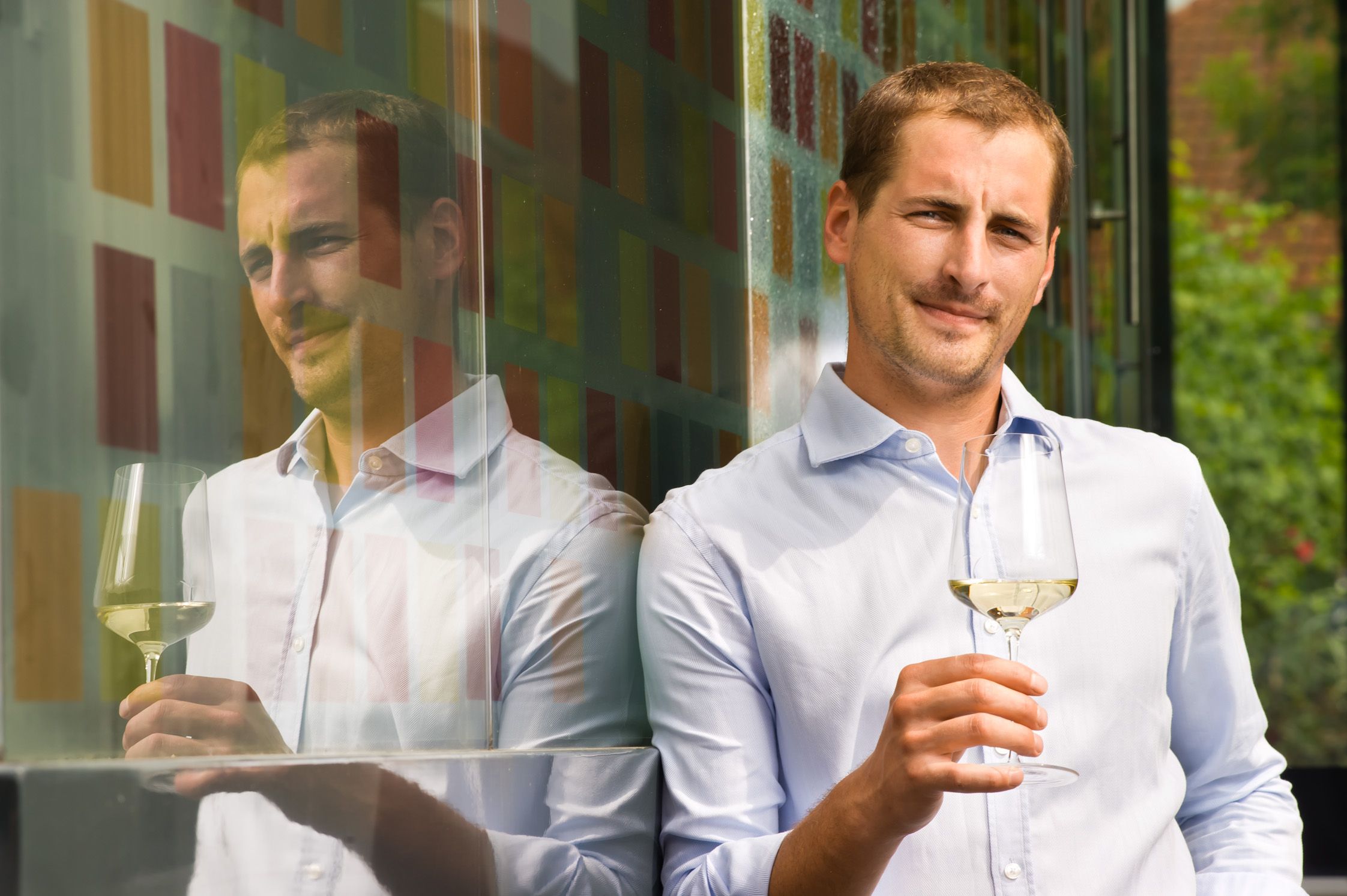 A man in a light blue shirt holds a glass of white wine and leans against a colorful glass wall.