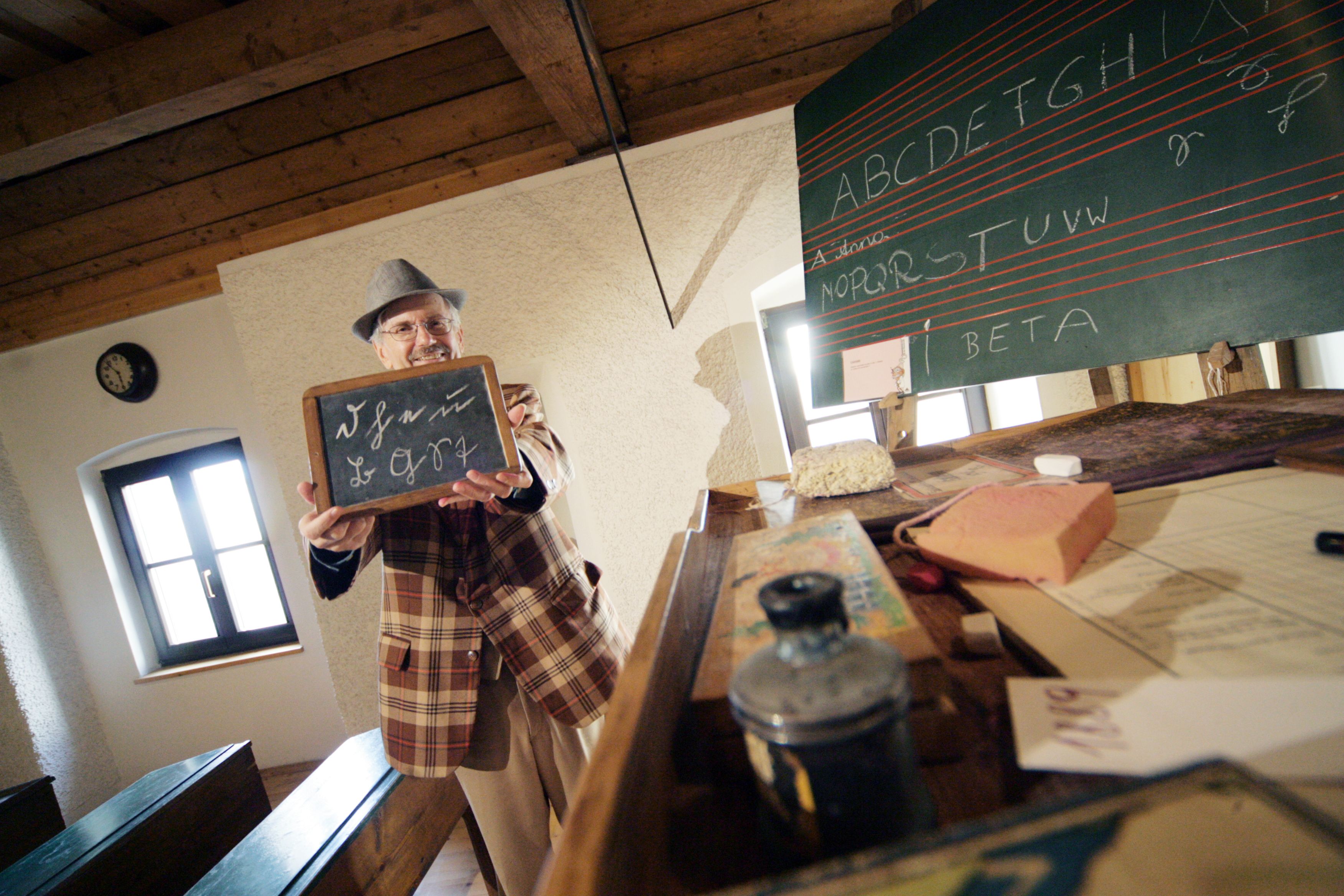 A man in a plaid suit holds a slate in a historic classroom.