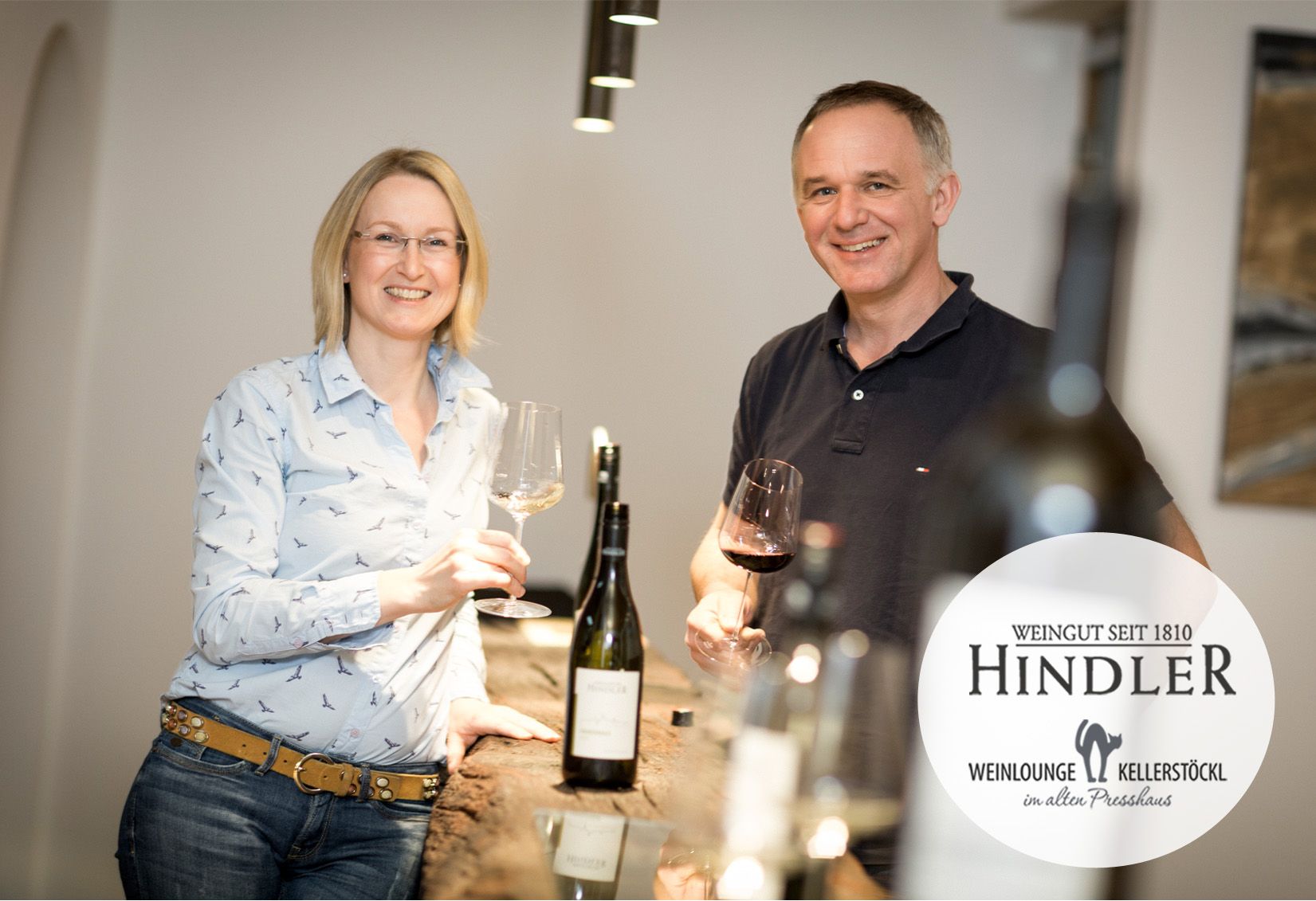 A pair of winemakers stand smiling with wine glasses in their hands at a table in the Hindler winery.