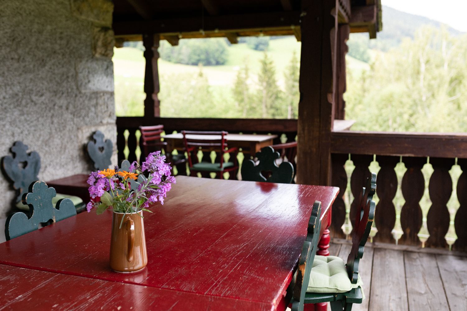 A rustic veranda with wooden tables and chairs, decorated with a jug full of colorful flowers. A green landscape can be seen in the background.