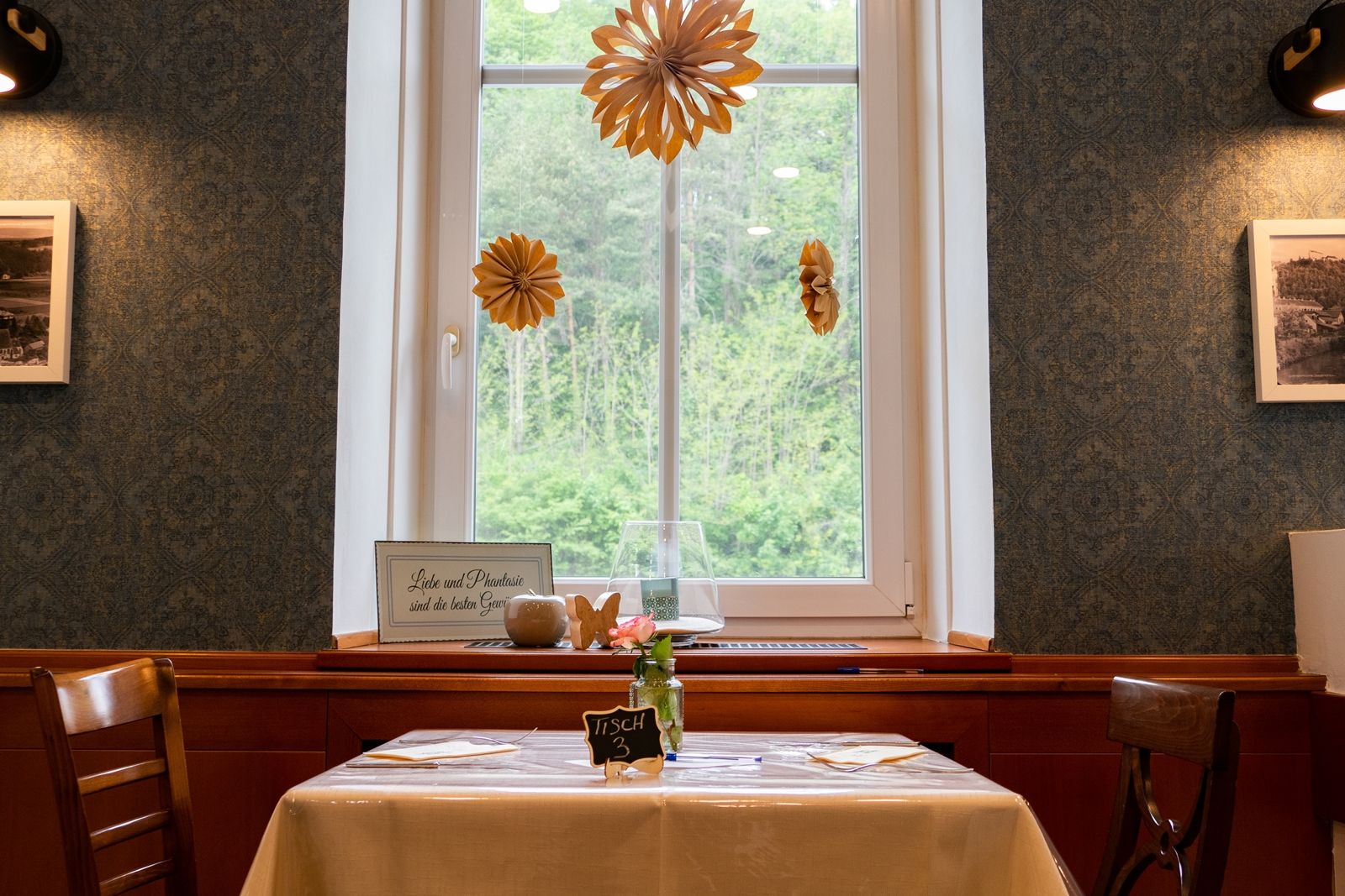 A table in a restaurant with a window view, decorated with paper flowers and a sign saying 'Love and imagination are the best spices'.