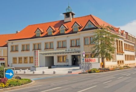 Traiskirchen town hall with red roof and several windows, surrounded by a street and flower beds.