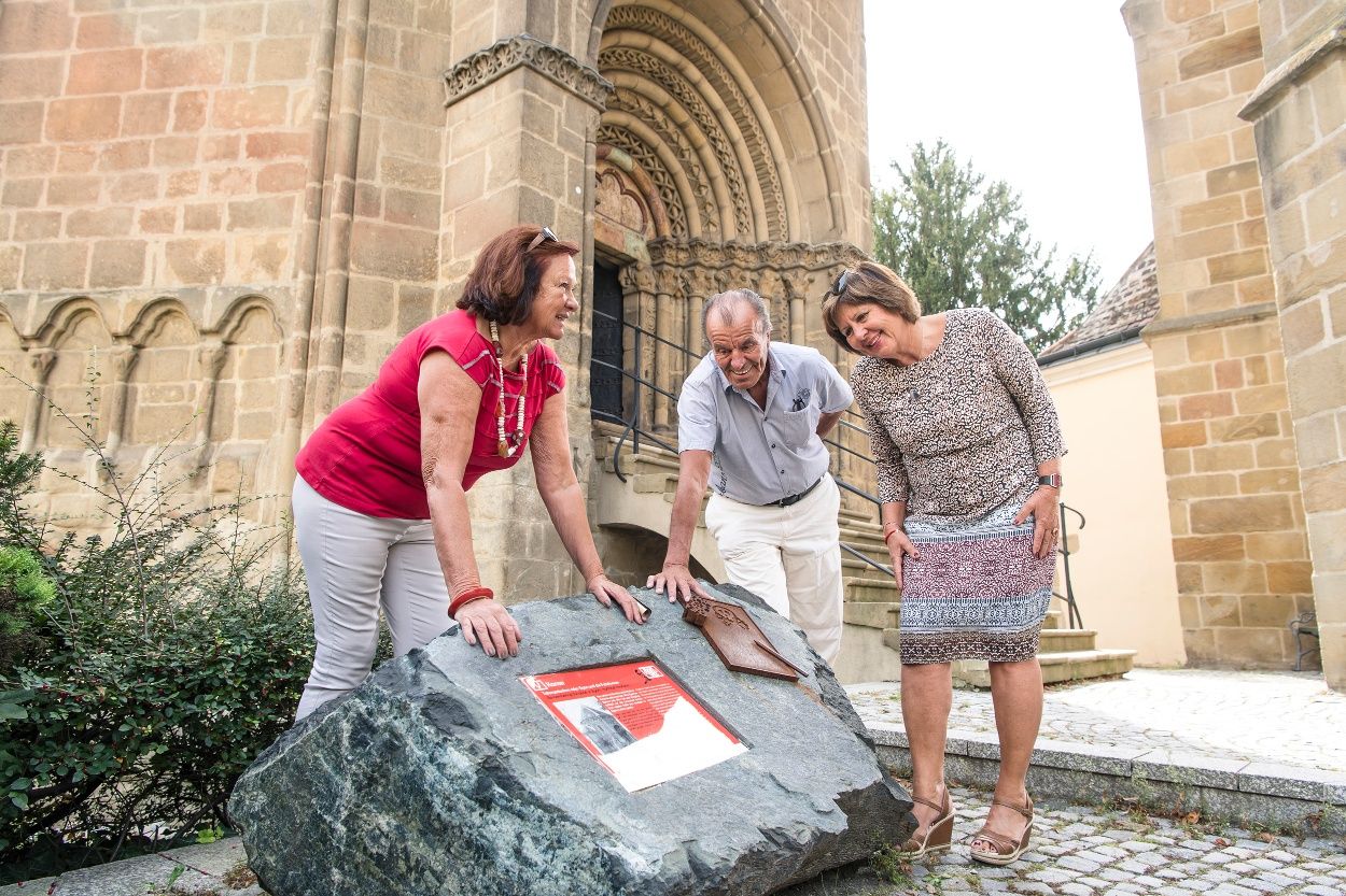 Three people look at a memorial plaque on a stone in front of a historic building.