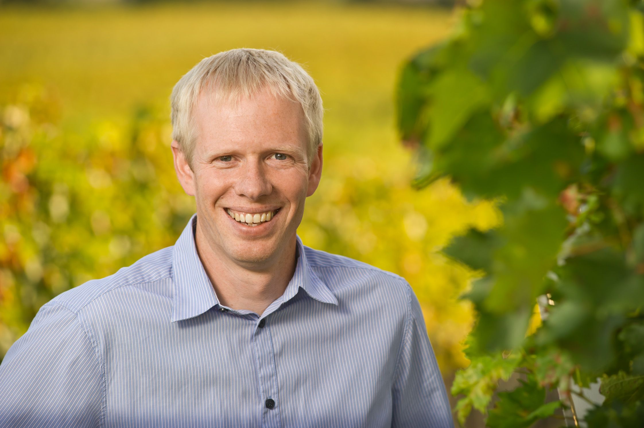 A smiling man in a blue shirt stands in front of a blurred background with green leaves.