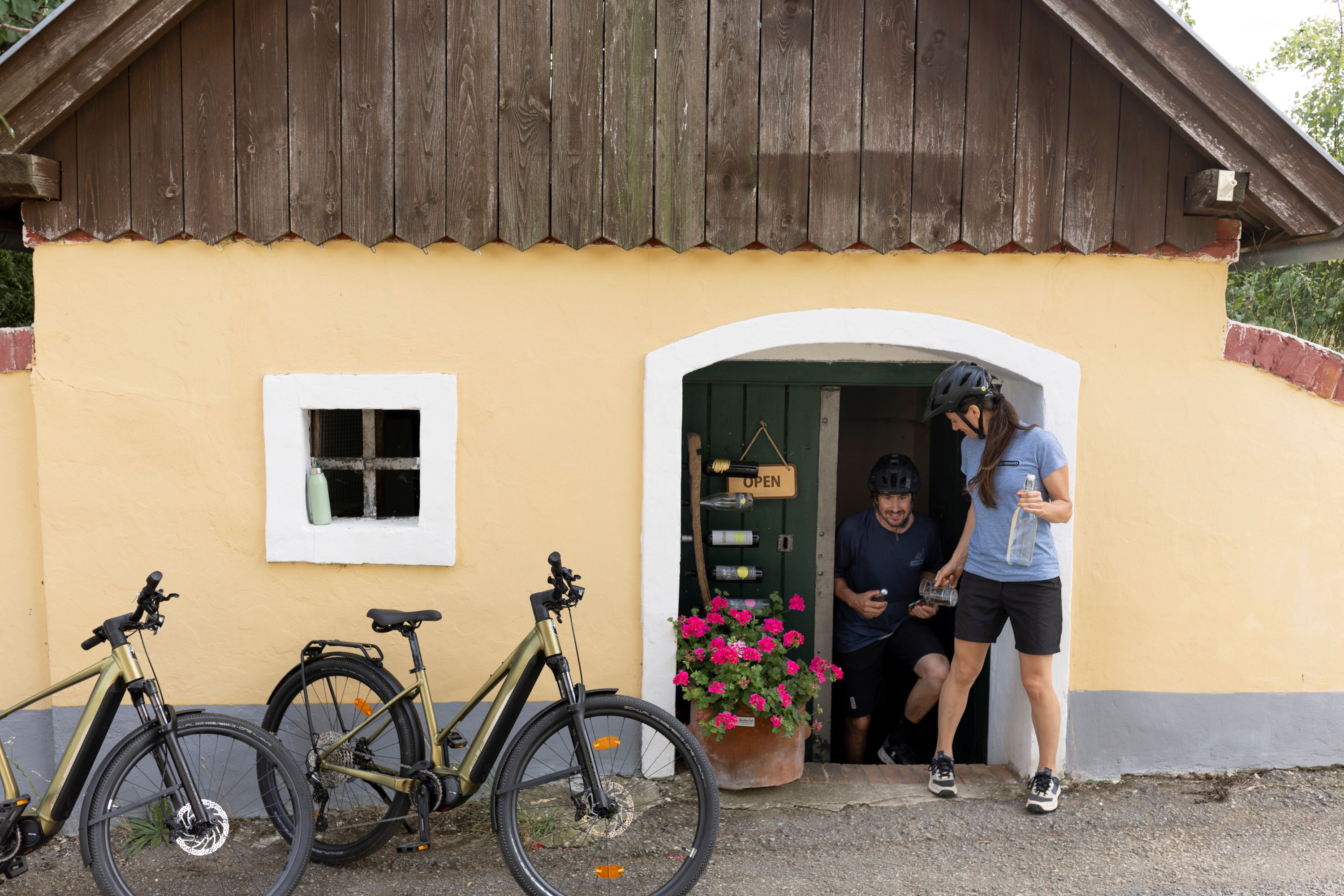 Two people with bicycles in front of a small yellow building with a wooden roof and flowers.