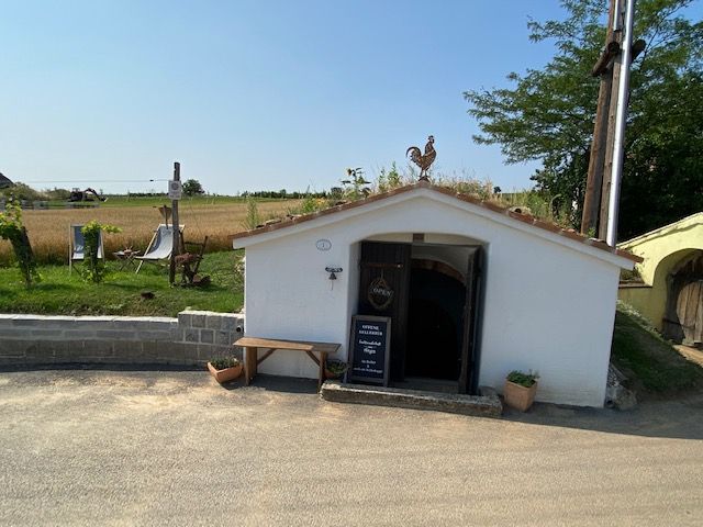 White cellar building with a rooster sculpture on the roof and an open entrance, surrounded by rural landscape.