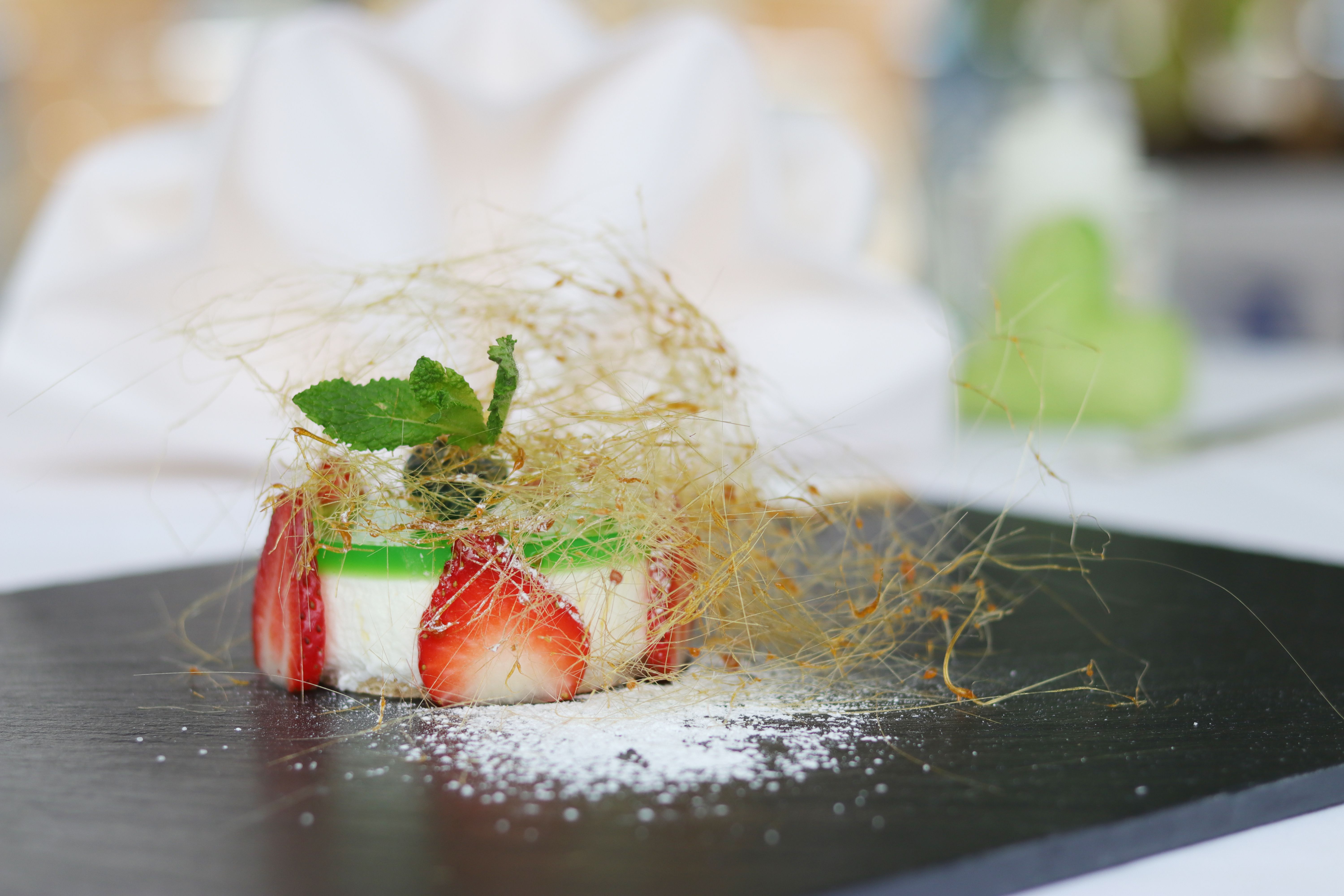 An artfully arranged goat's cheese tartlet with strawberries and caramel threads on a black plate.