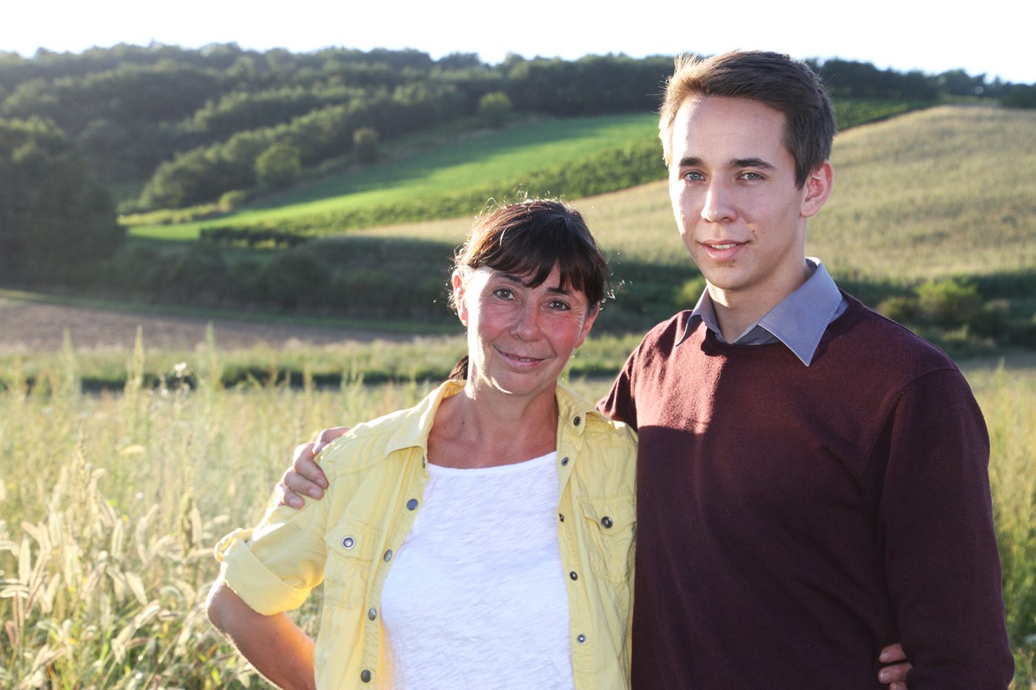 A woman and a young man stand in a rural landscape and smile at the camera.