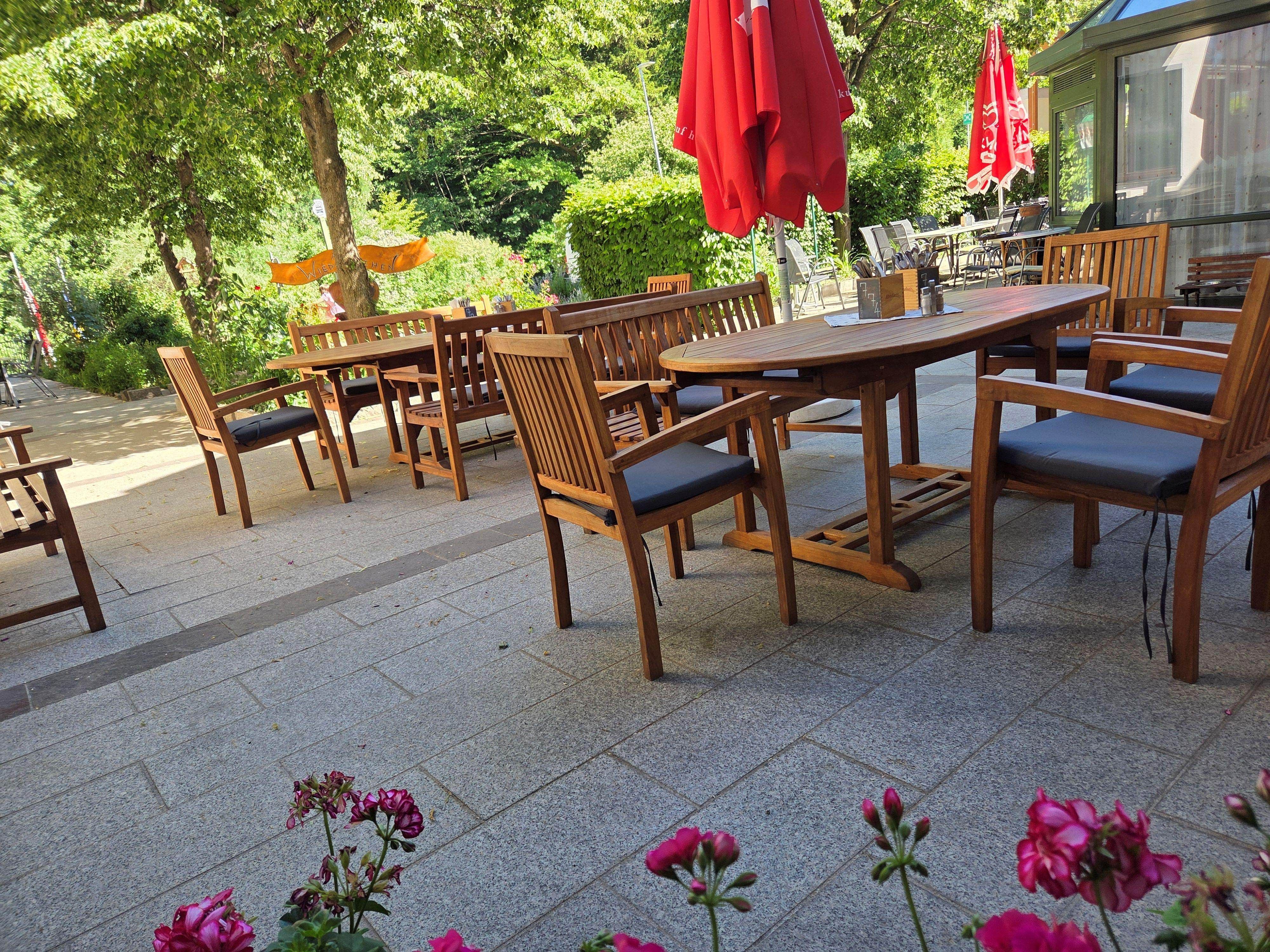 A cozy outdoor dining area with wooden furniture and red umbrellas, surrounded by green trees and flowers.