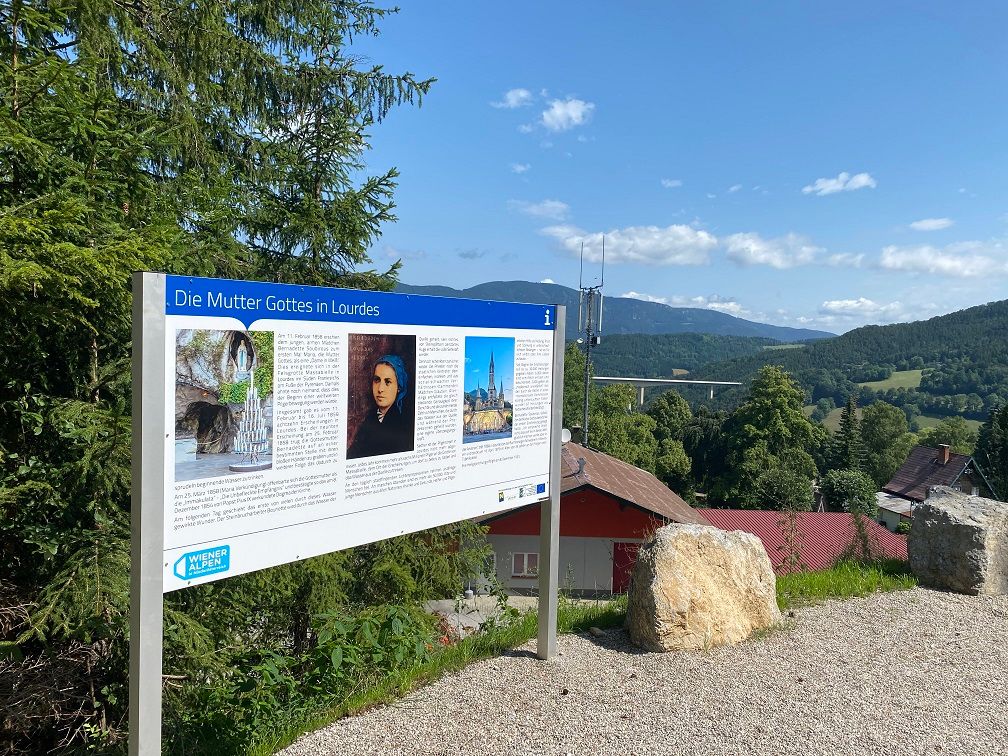 Information sign about the Mother of God in Lourdes in front of a landscape with hills and trees.