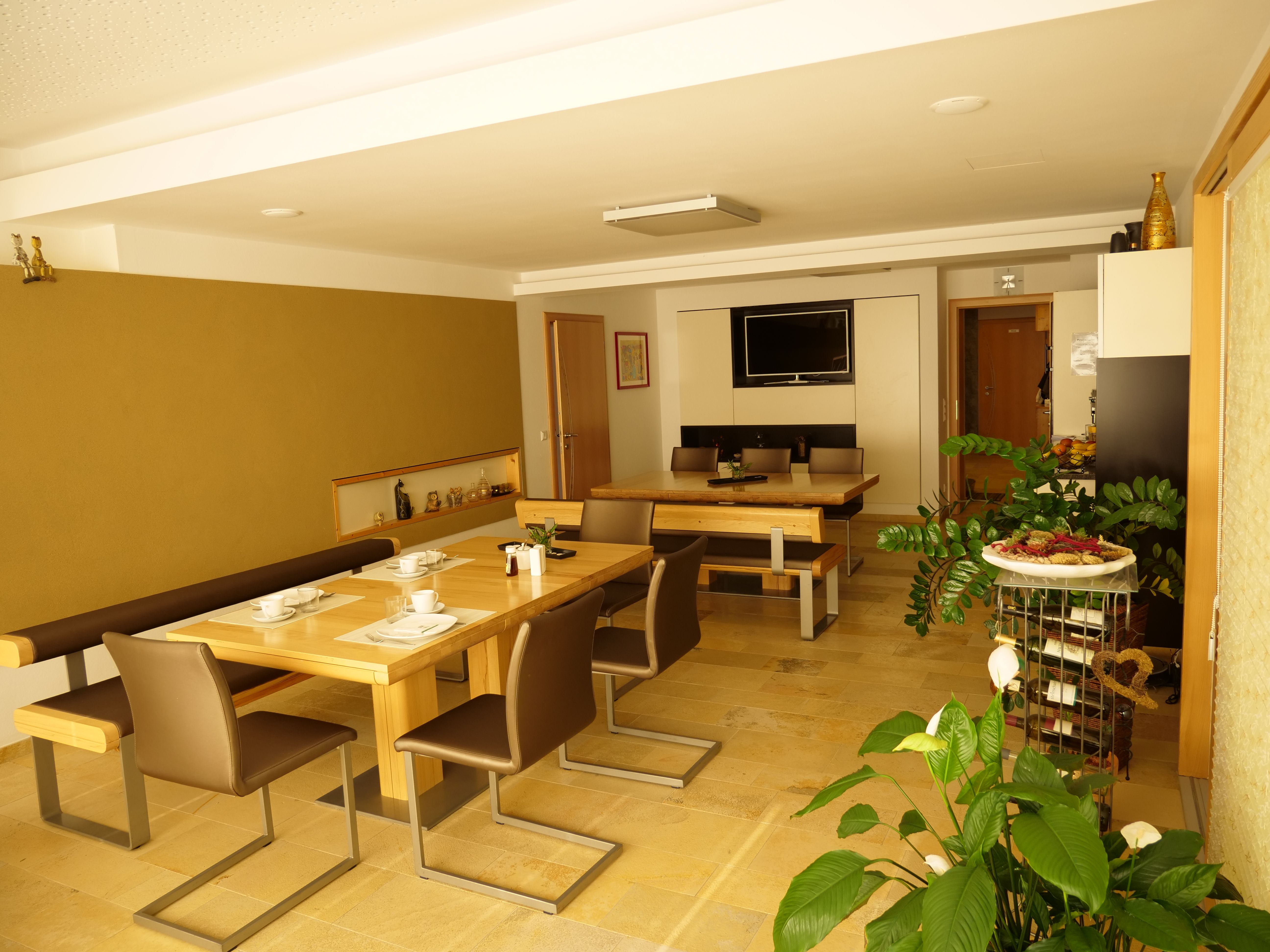 A breakfast room with wooden tables, chairs and plants, decorated with crockery and a TV on the wall.