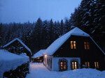 Snow-covered house in the forest at dusk with illuminated windows.