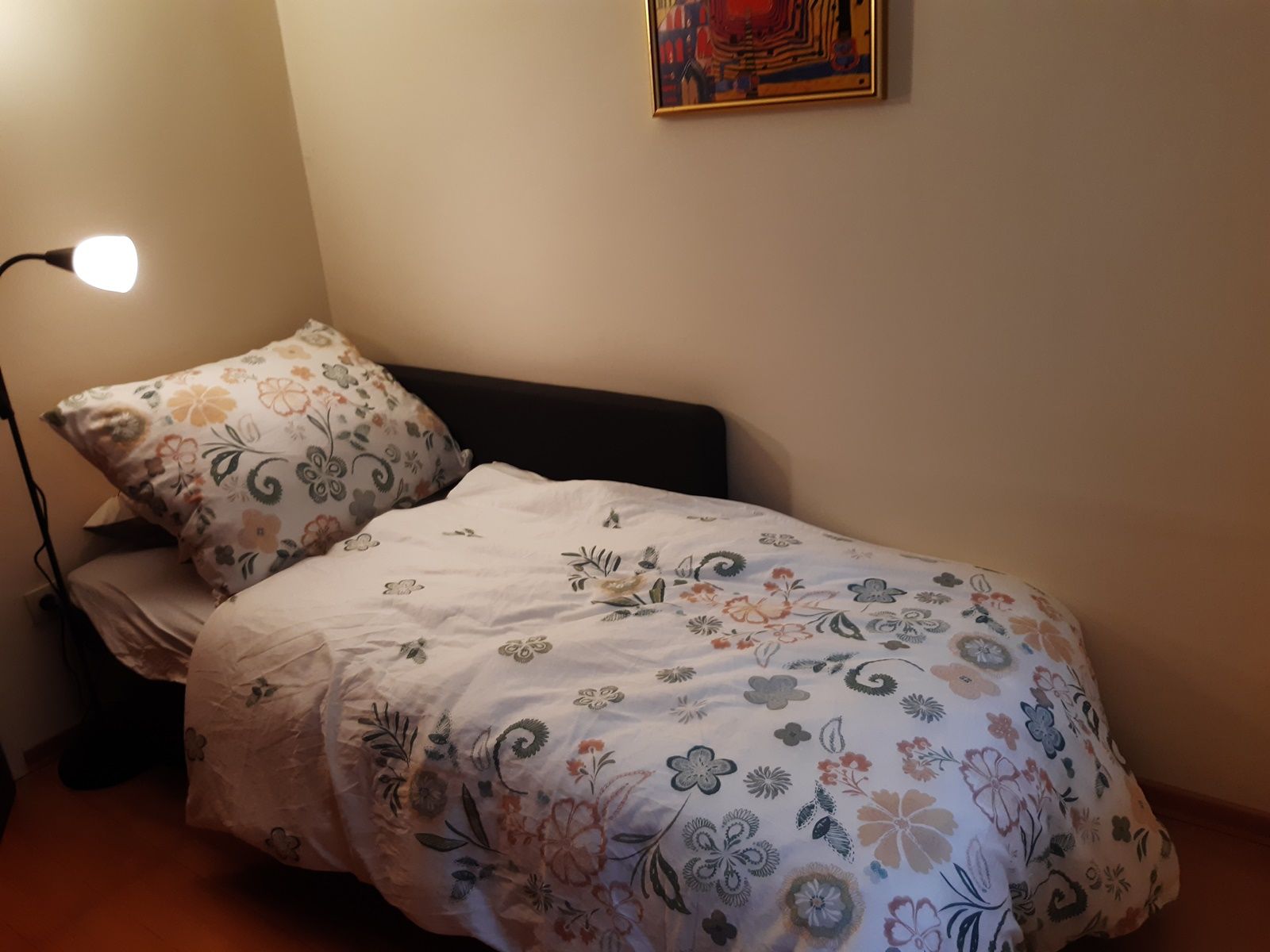 A small room with a single bed covered with a flowered bedspread. A floor lamp stands next to it and a colorful picture hangs on the wall.