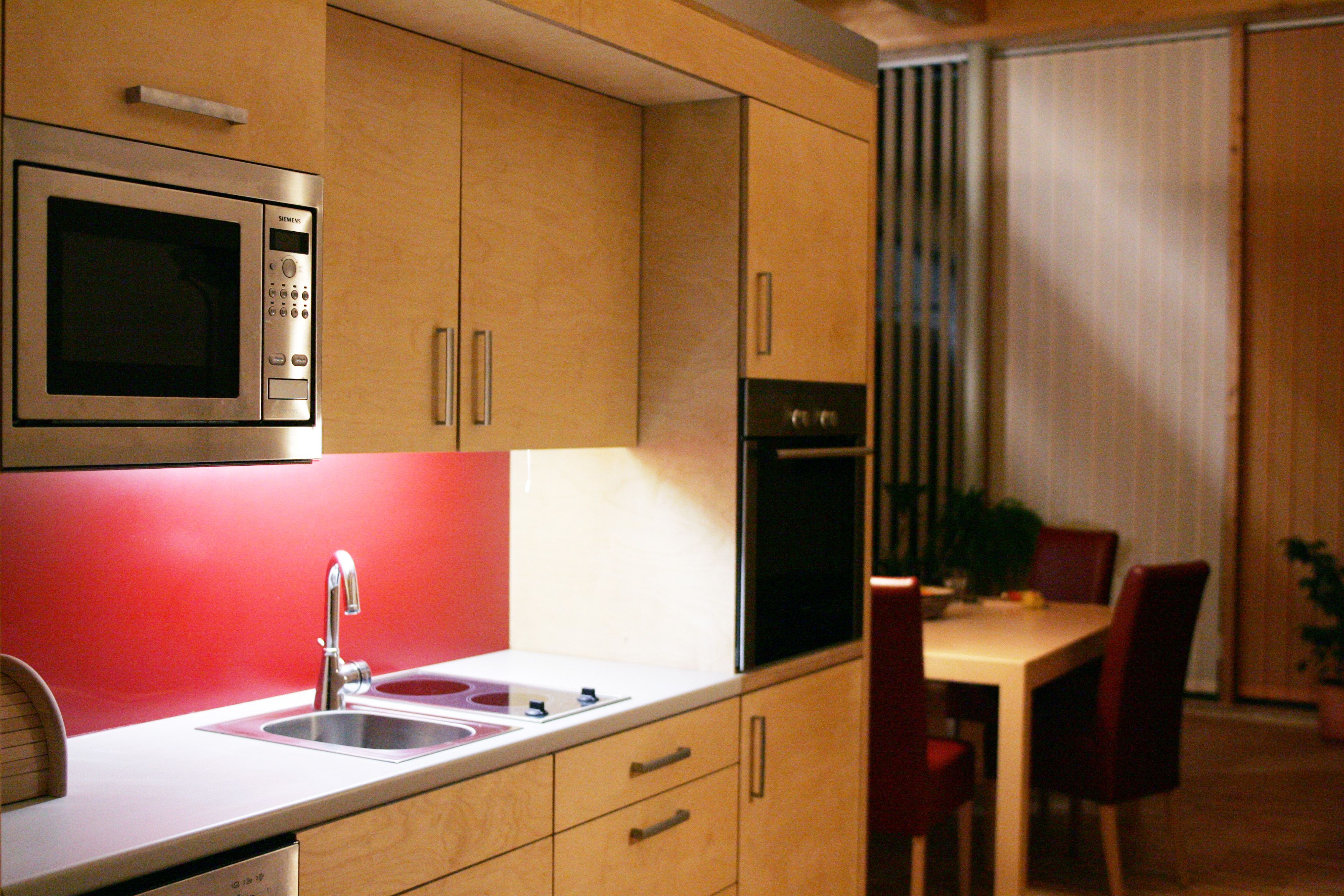 Modern kitchen with wooden cupboards, red splashback, microwave and dining table in the background.