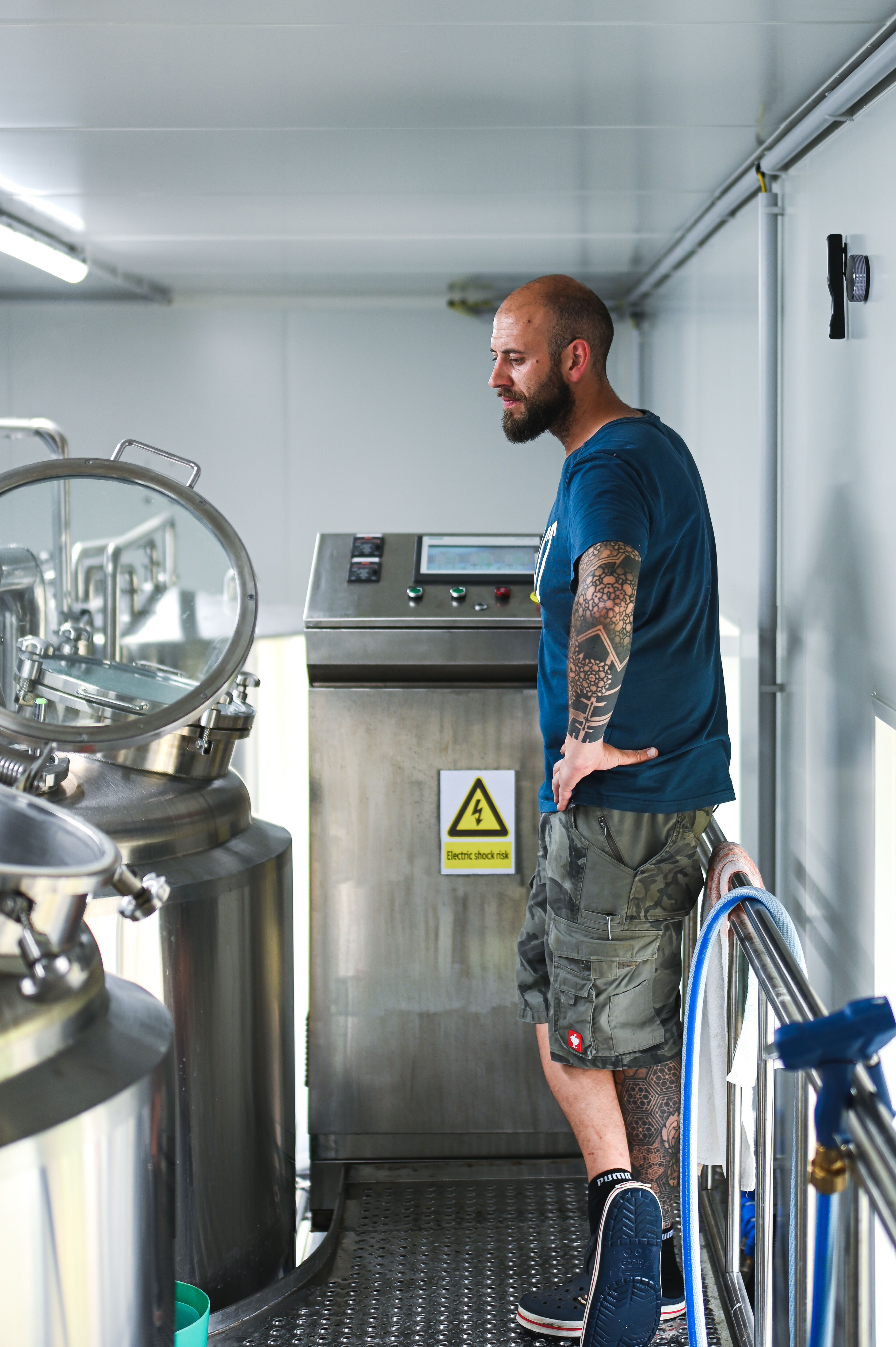 A man with a tattooed arm stands in a brewery next to a stainless steel container and a control console.