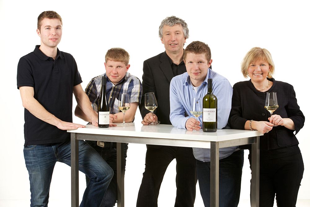 Five people are standing around a table with bottles of wine and glasses.
