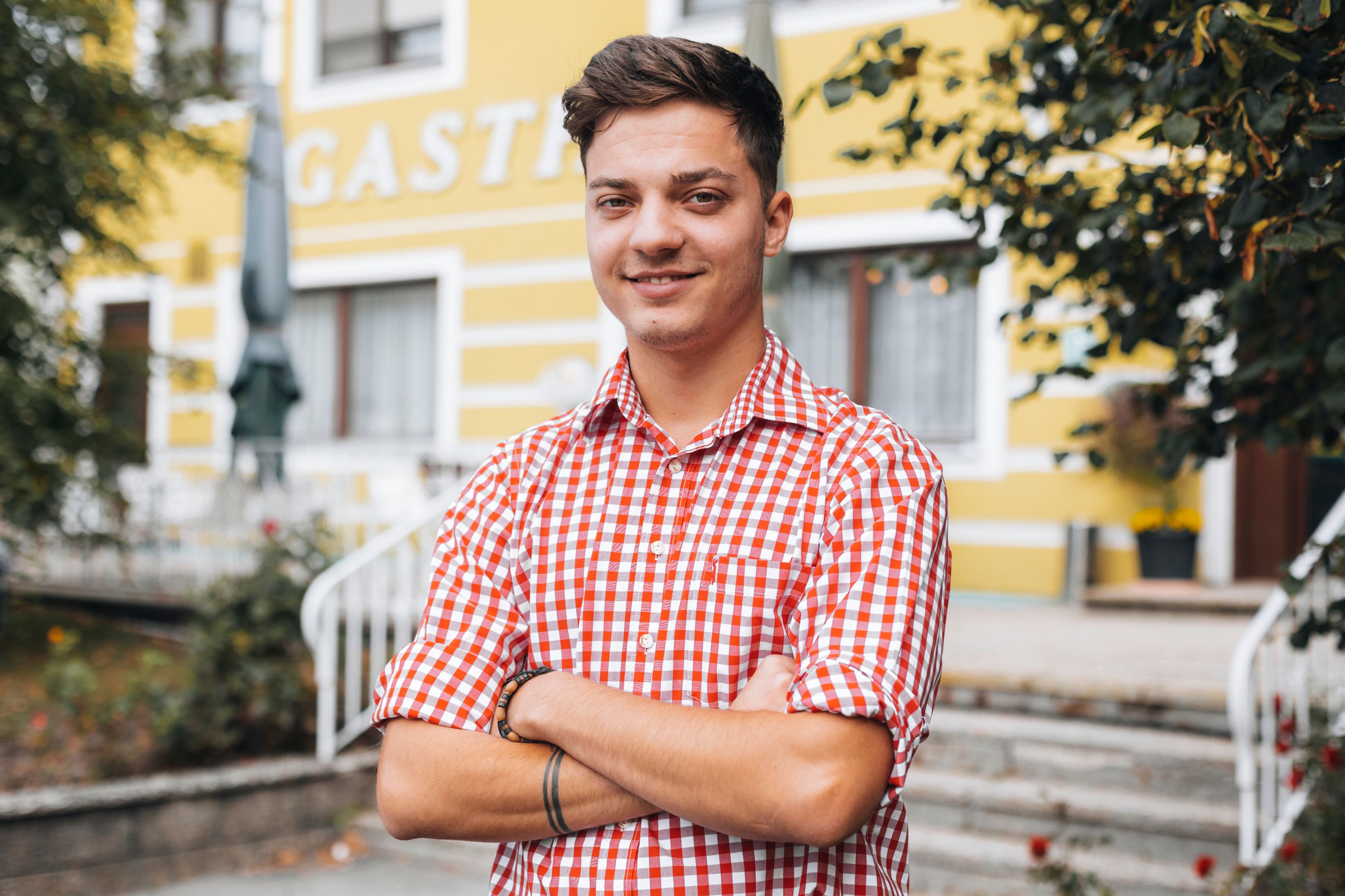 A young man in a checked shirt stands smiling in front of a yellow building.